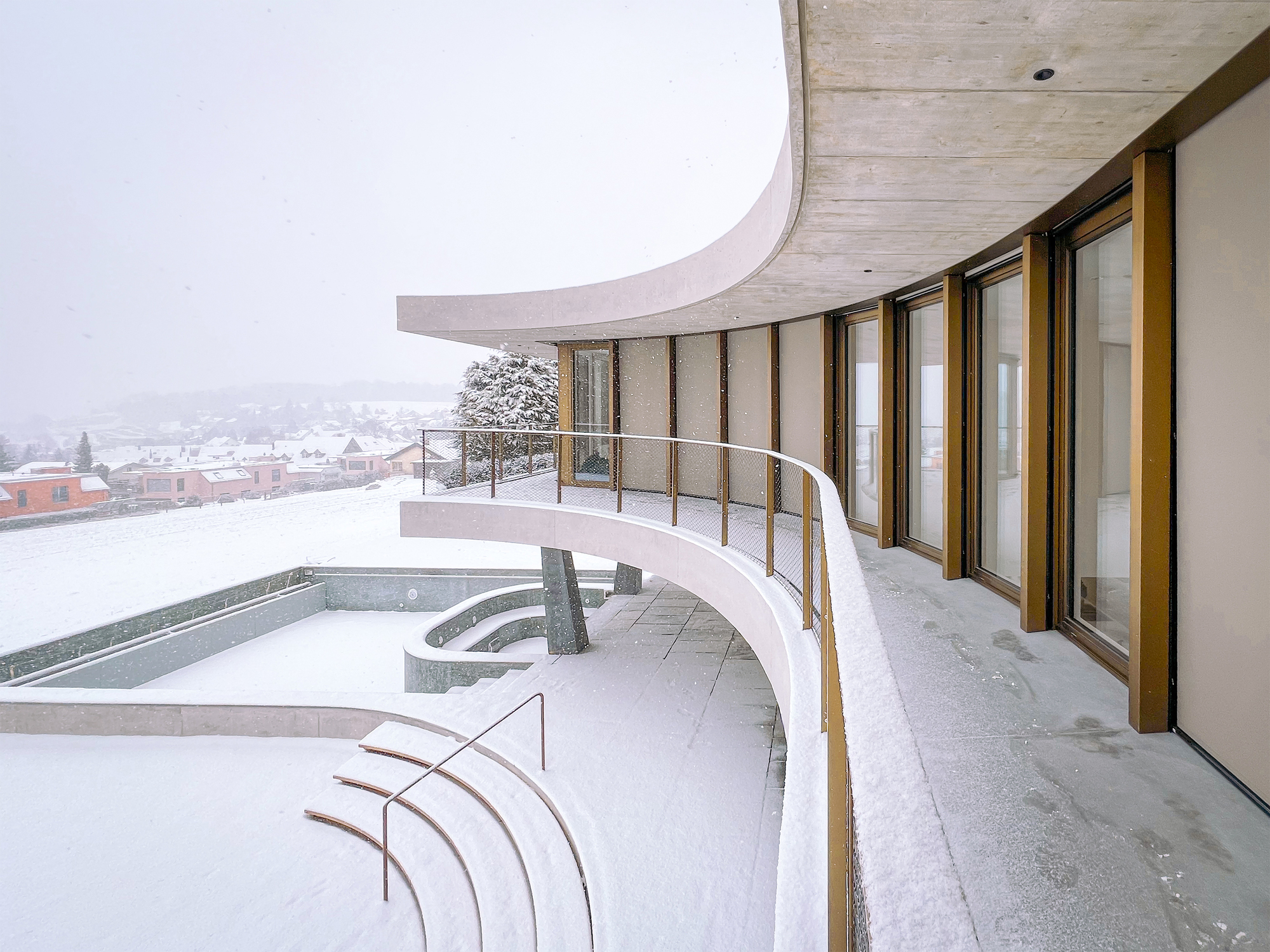 Curved terrace with timber soffit and mesh railing overlooking snow-covered rooftops and hills