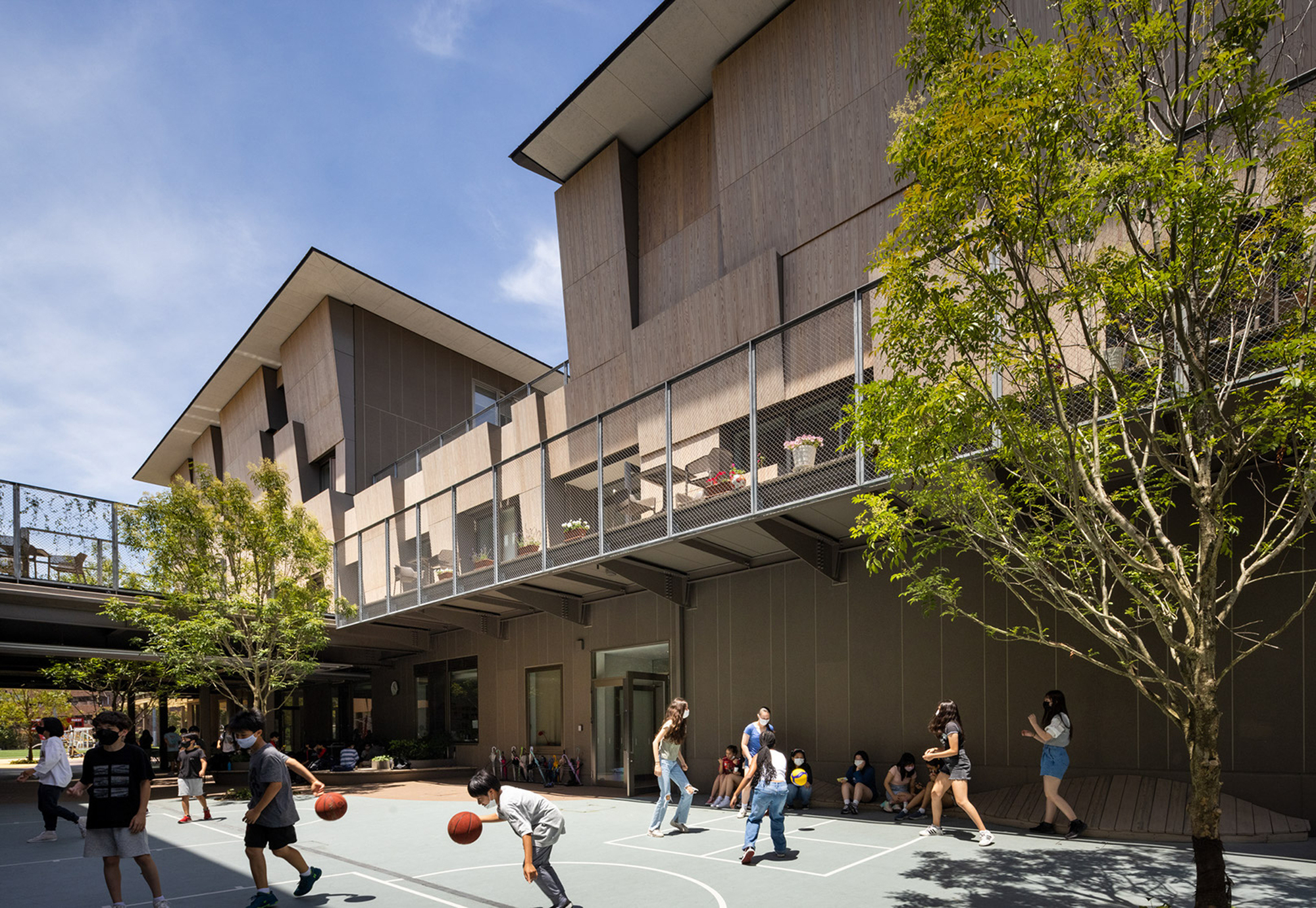 Outdoor basketball court surrounded by timber-clad volumes with an elevated pedestrian bridge and spring foliage