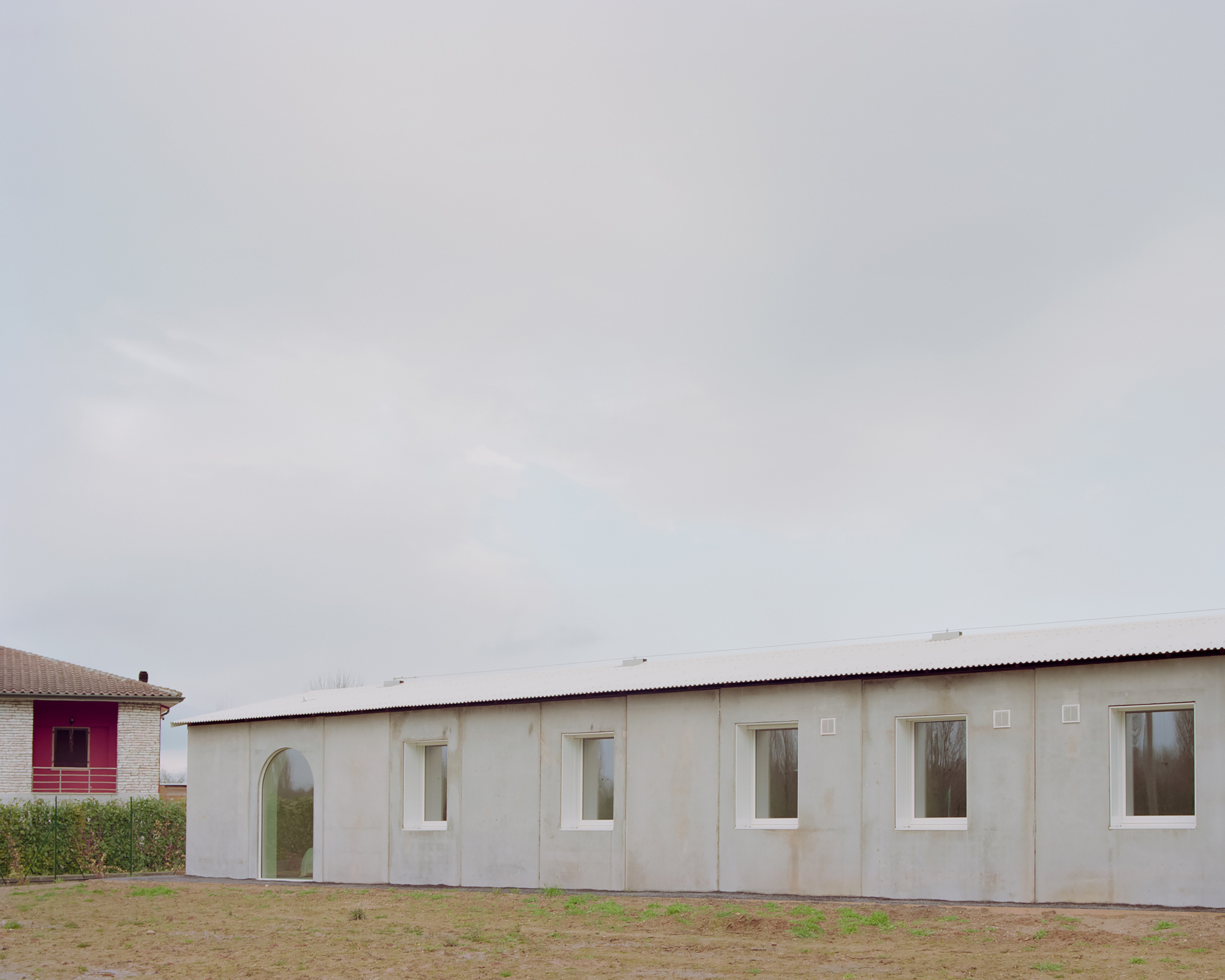 Pale plaster facade with rhythmic rectangular windows and corrugated metal roof under overcast sky