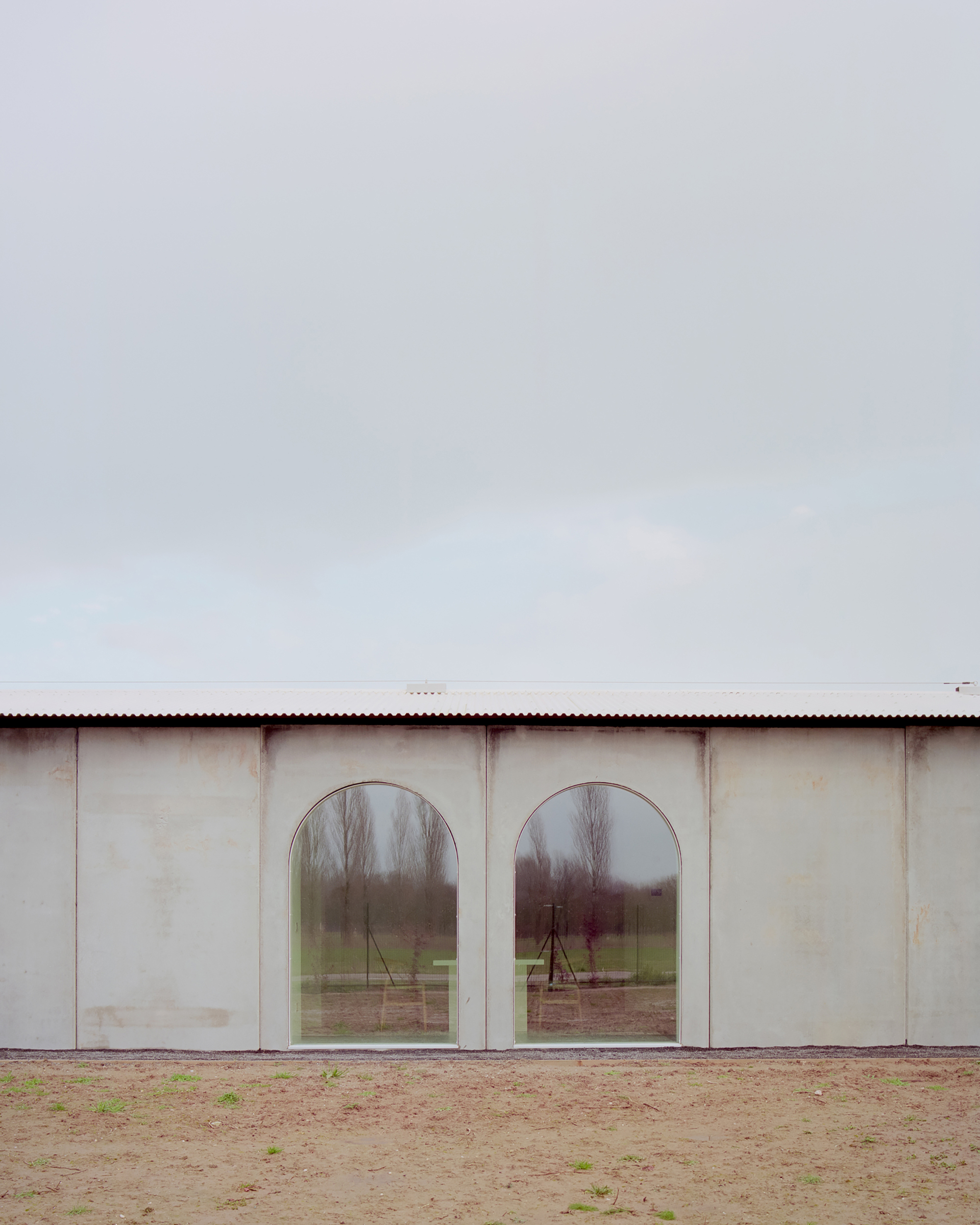 Twin arched windows in plaster facade reflecting bare trees and playground beyond