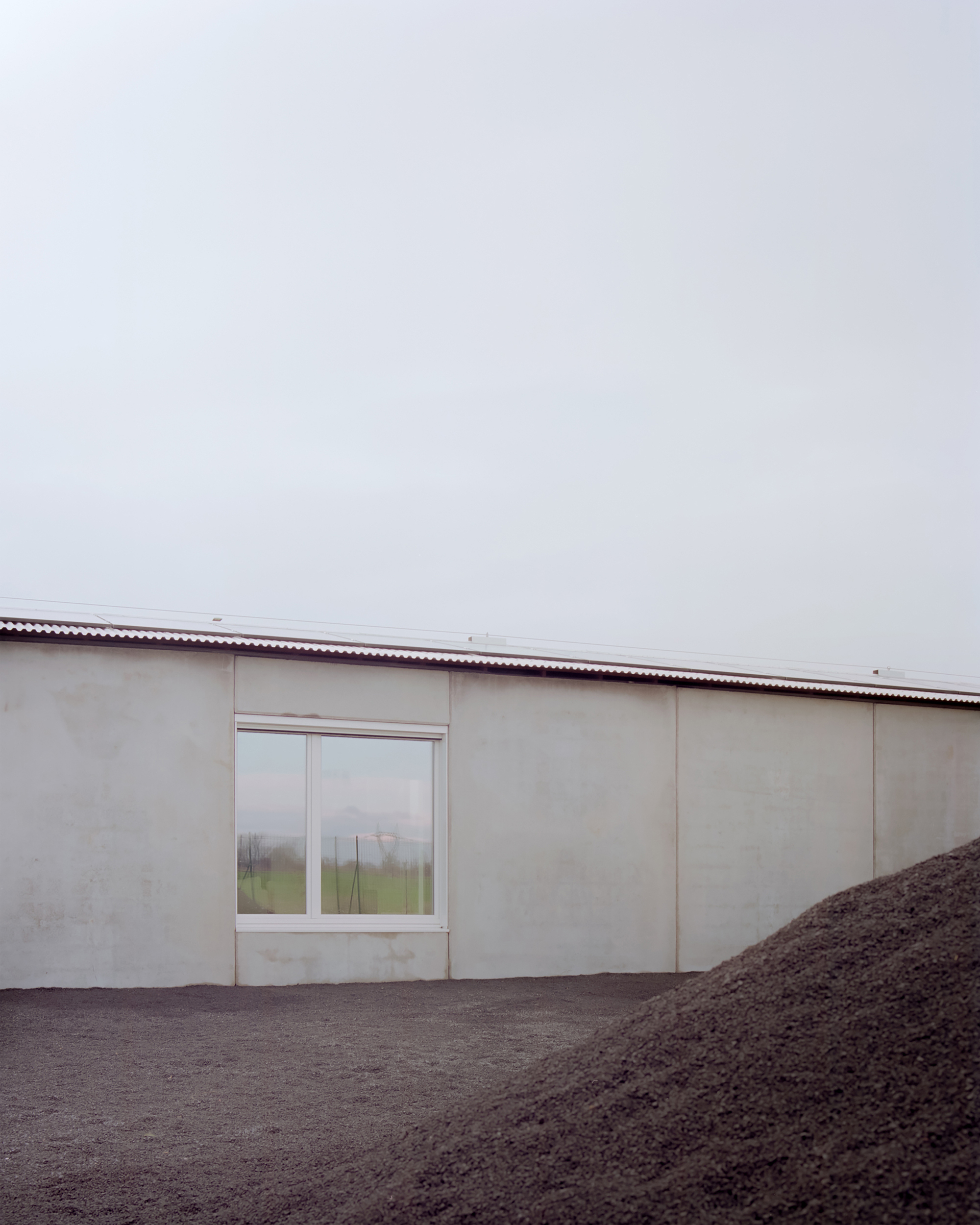 Pale facade with single window and corrugated metal roof beside mound of dark soil