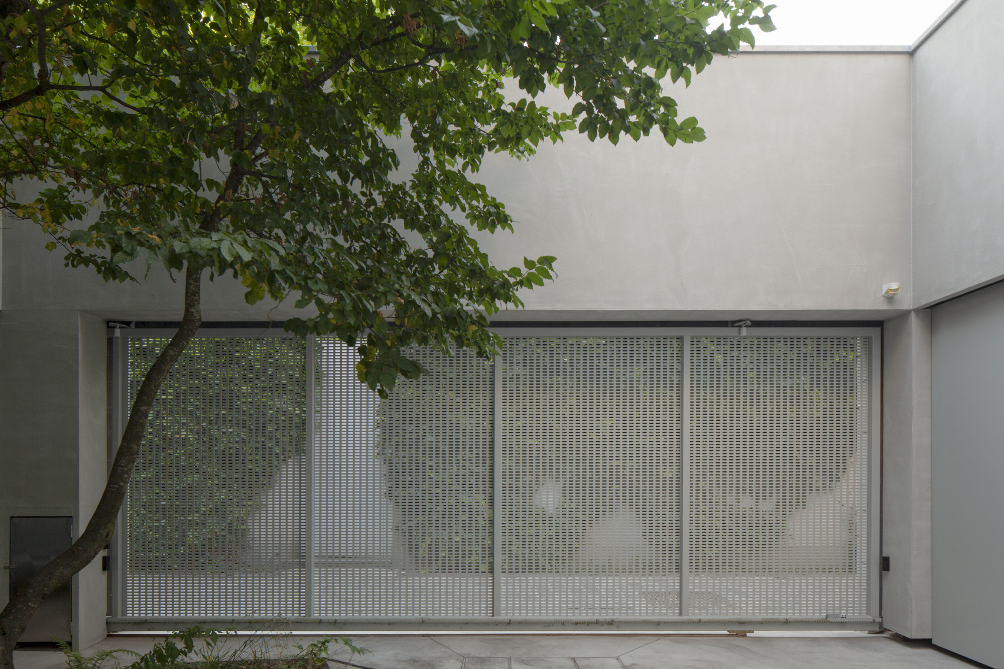 Perforated metal gate beneath tree canopy framed by rendered concrete walls