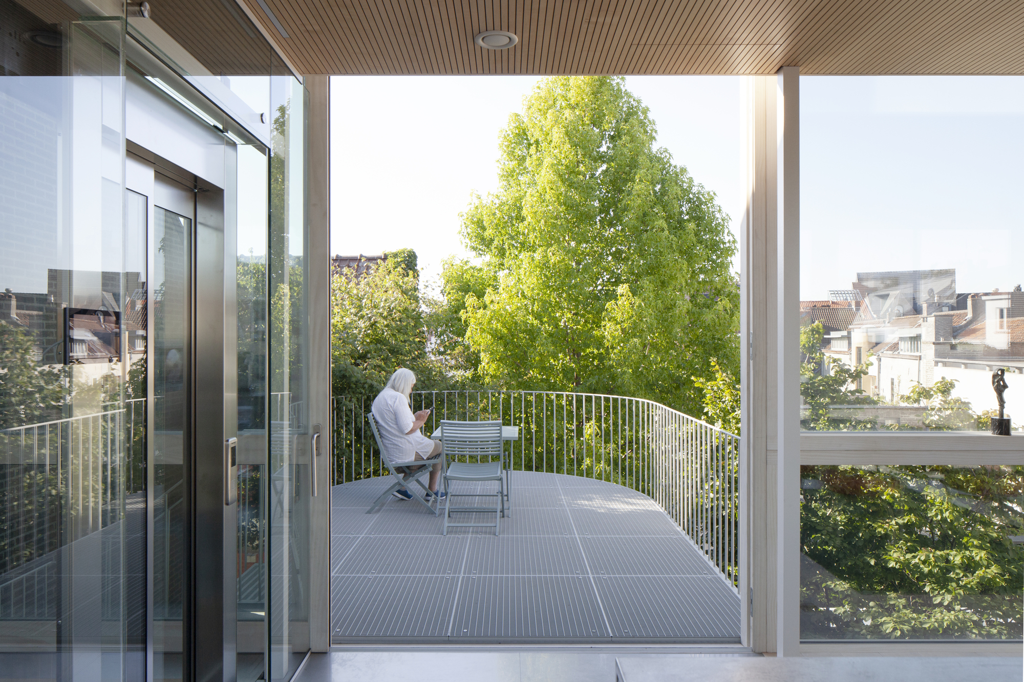 Covered terrace with slatted timber ceiling where a person sits reading beside tree