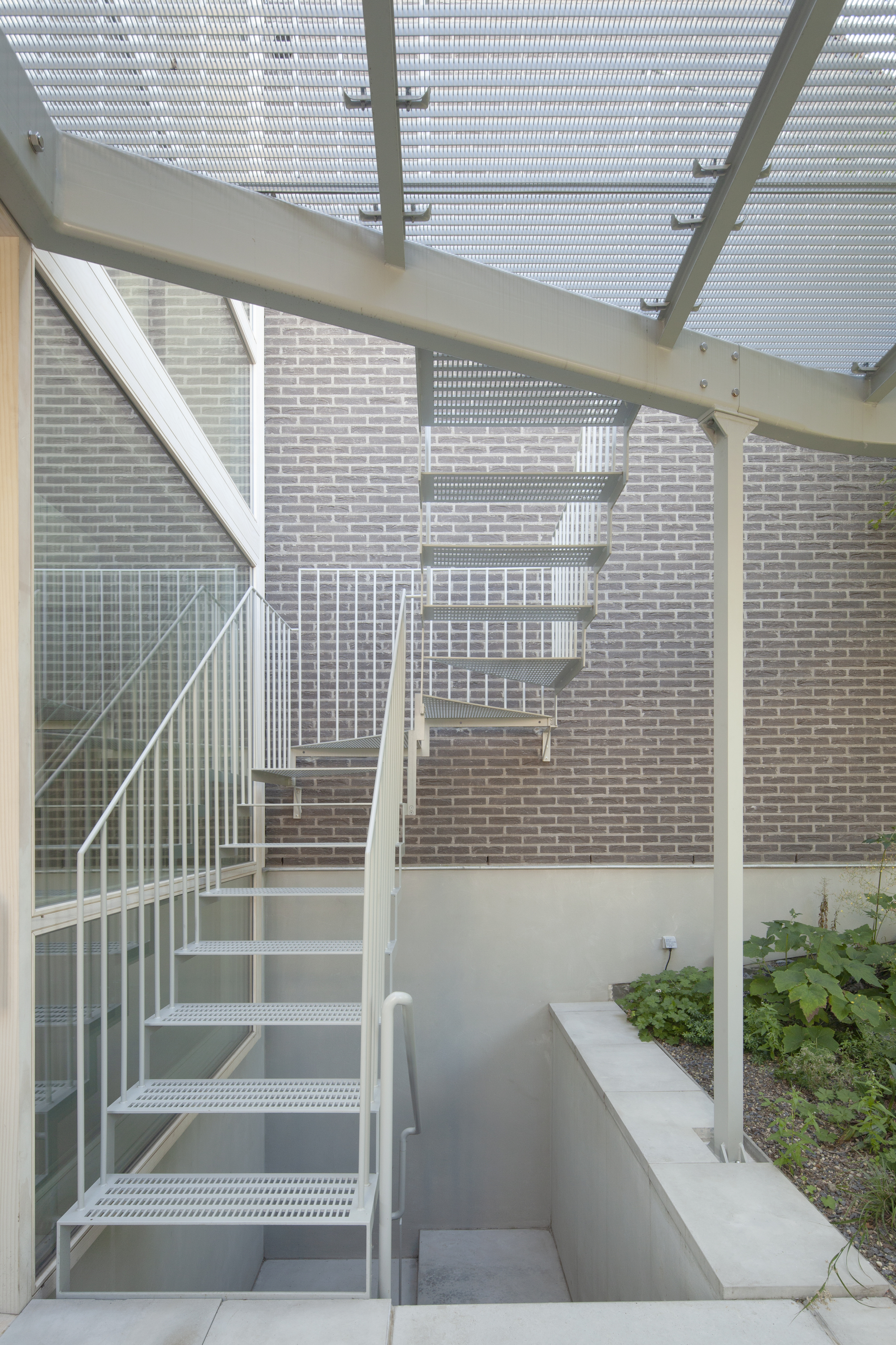 White perforated metal stair with railing ascending alongside a pale brick wall under a slatted pergola