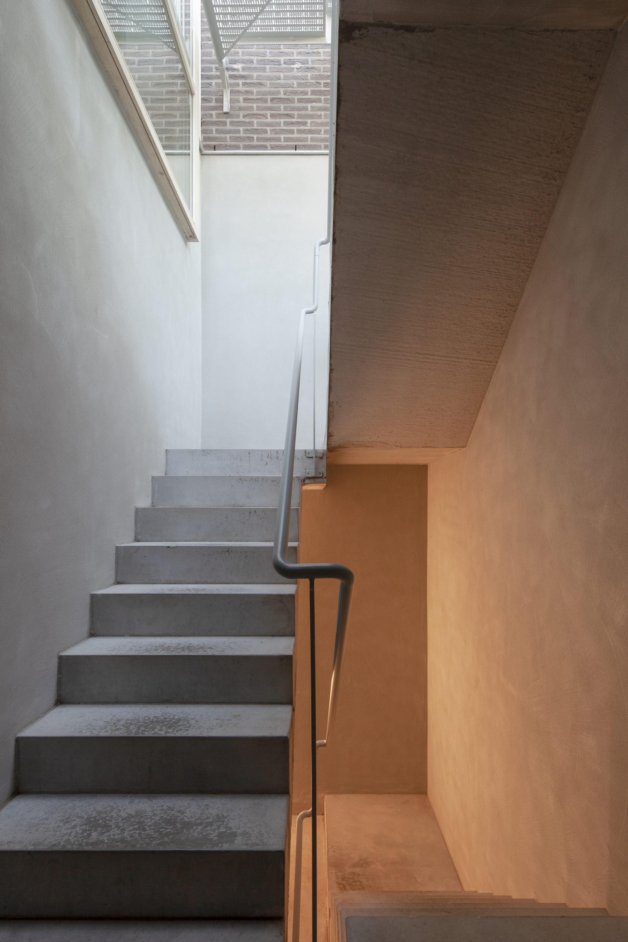 Concrete stair with black metal handrail ascending to glazed opening framing brick wall beyond