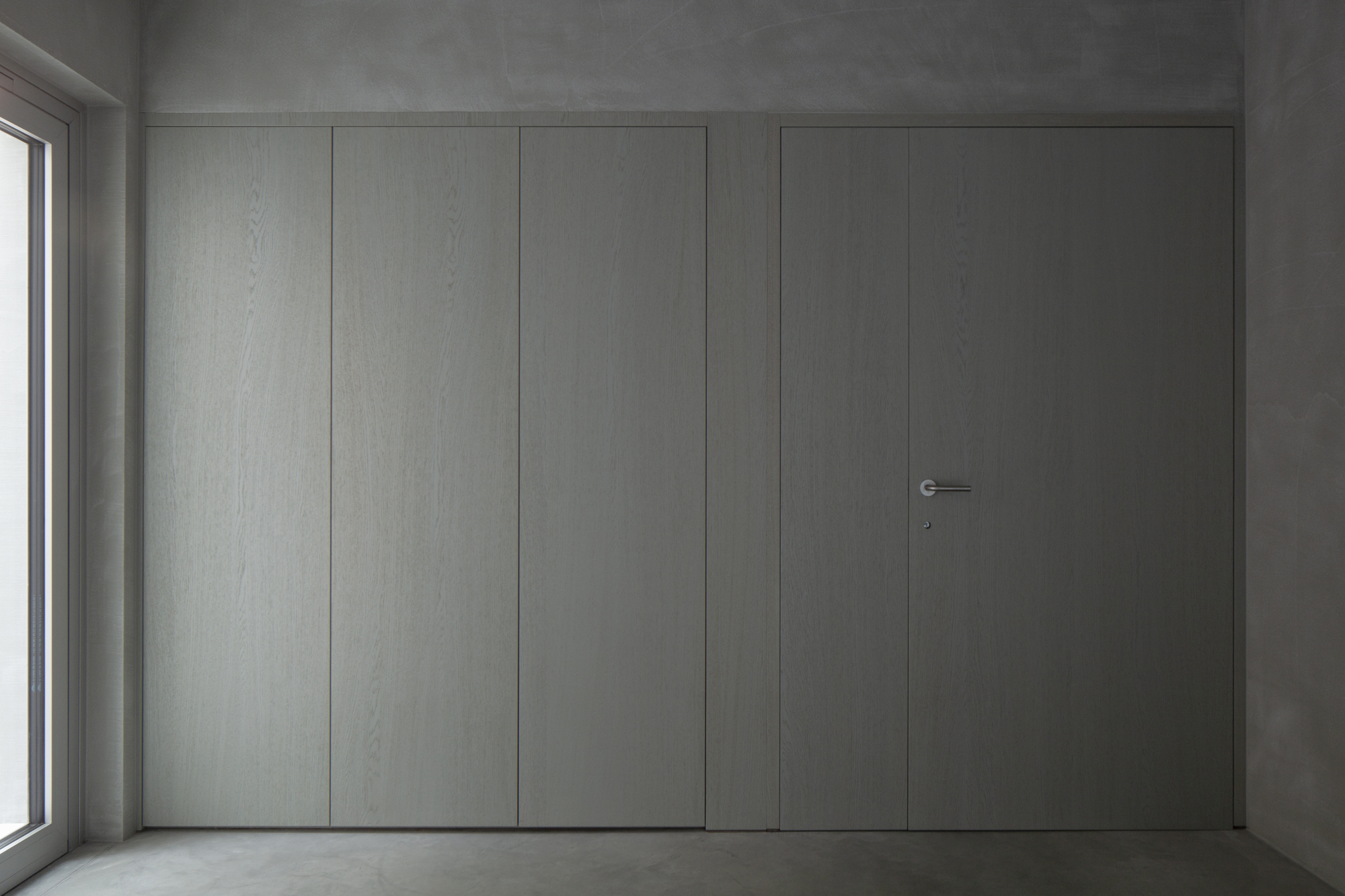 Flush pale grey paneled wall with concealed door and minimal hardware beside a window