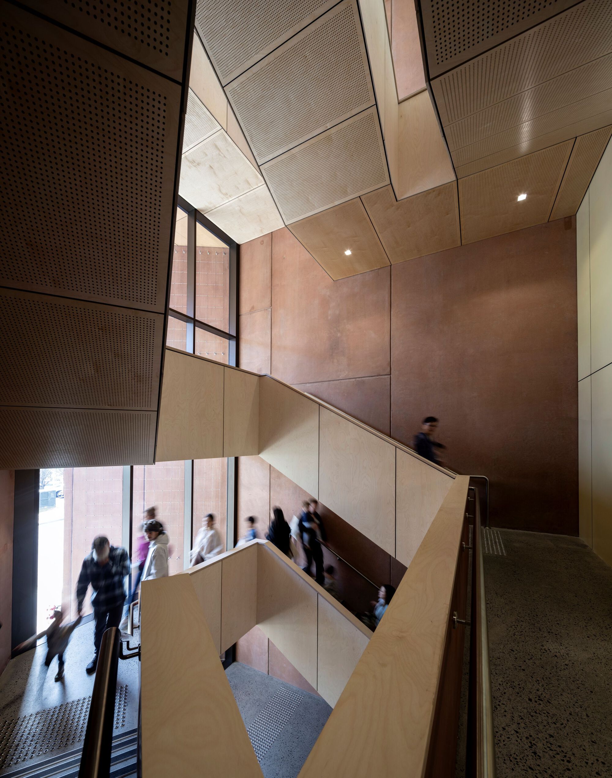Angular staircase with timber-paneled walls and perforated ceiling panels as visitors ascend between floors
