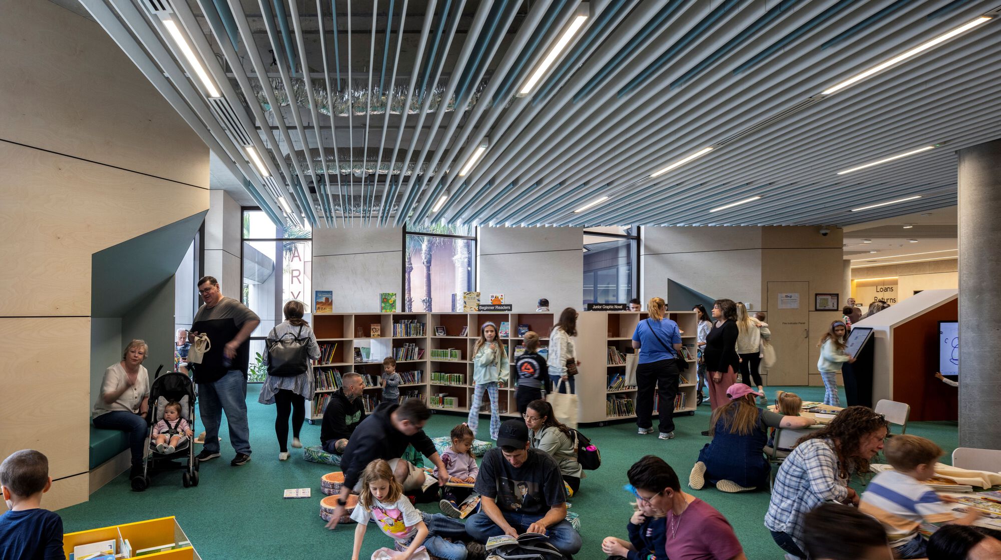 Library space with linear metal ceiling baffles and children seated on green carpet reading