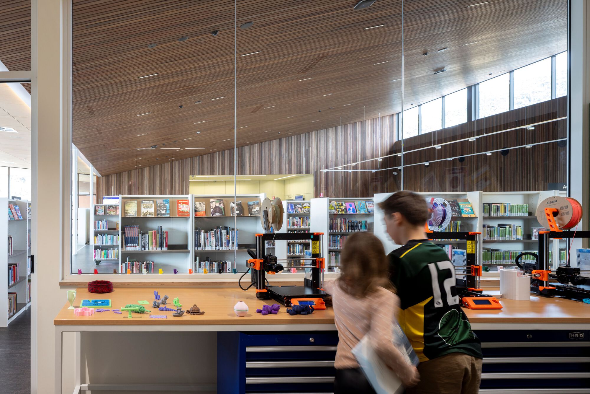 Makerspace workbench with 3D printers and blurred figures moving through the timber-clad room