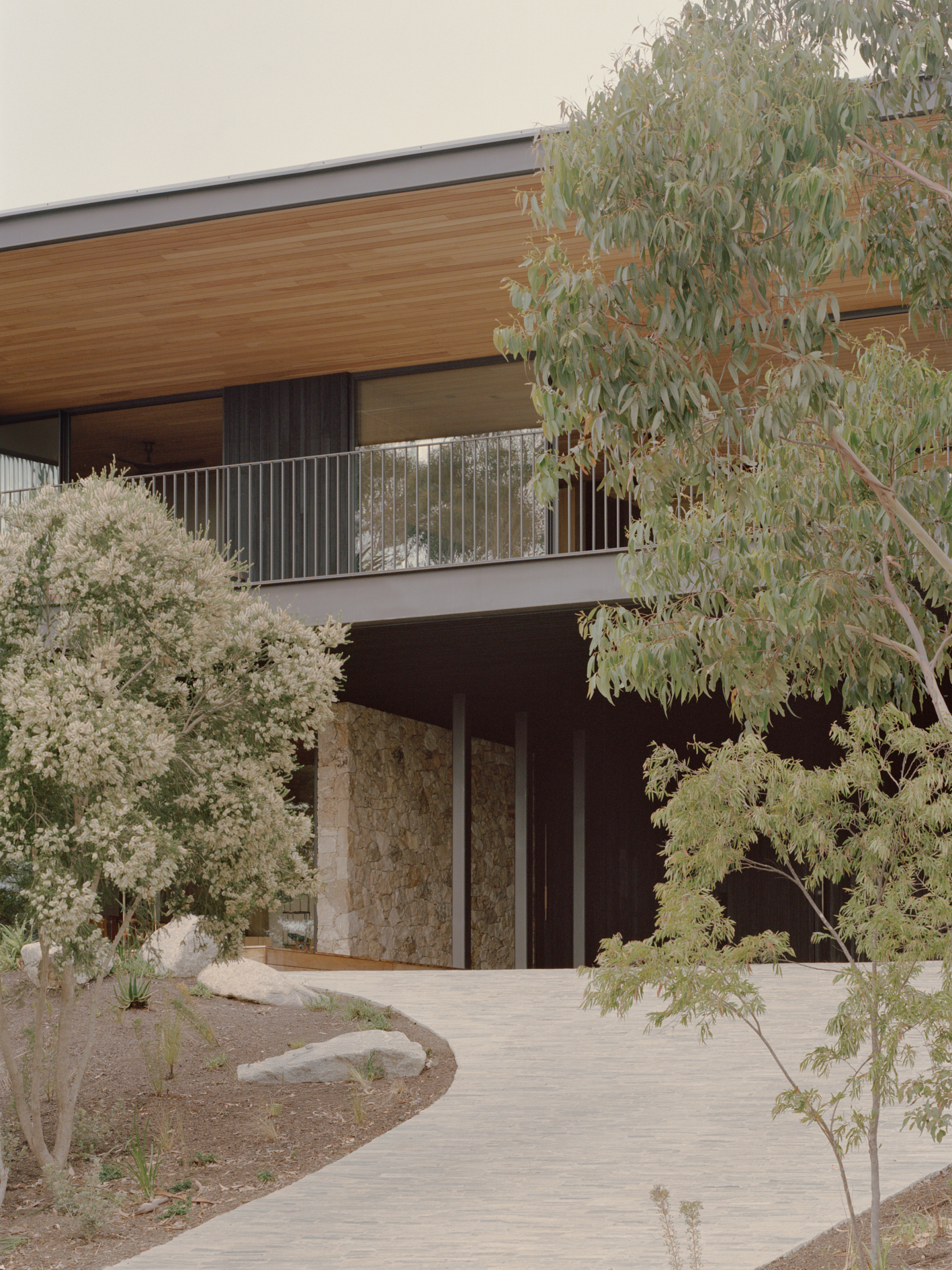 Entry facade with stone-clad walls and timber soffit framed by native plantings and curved pathway