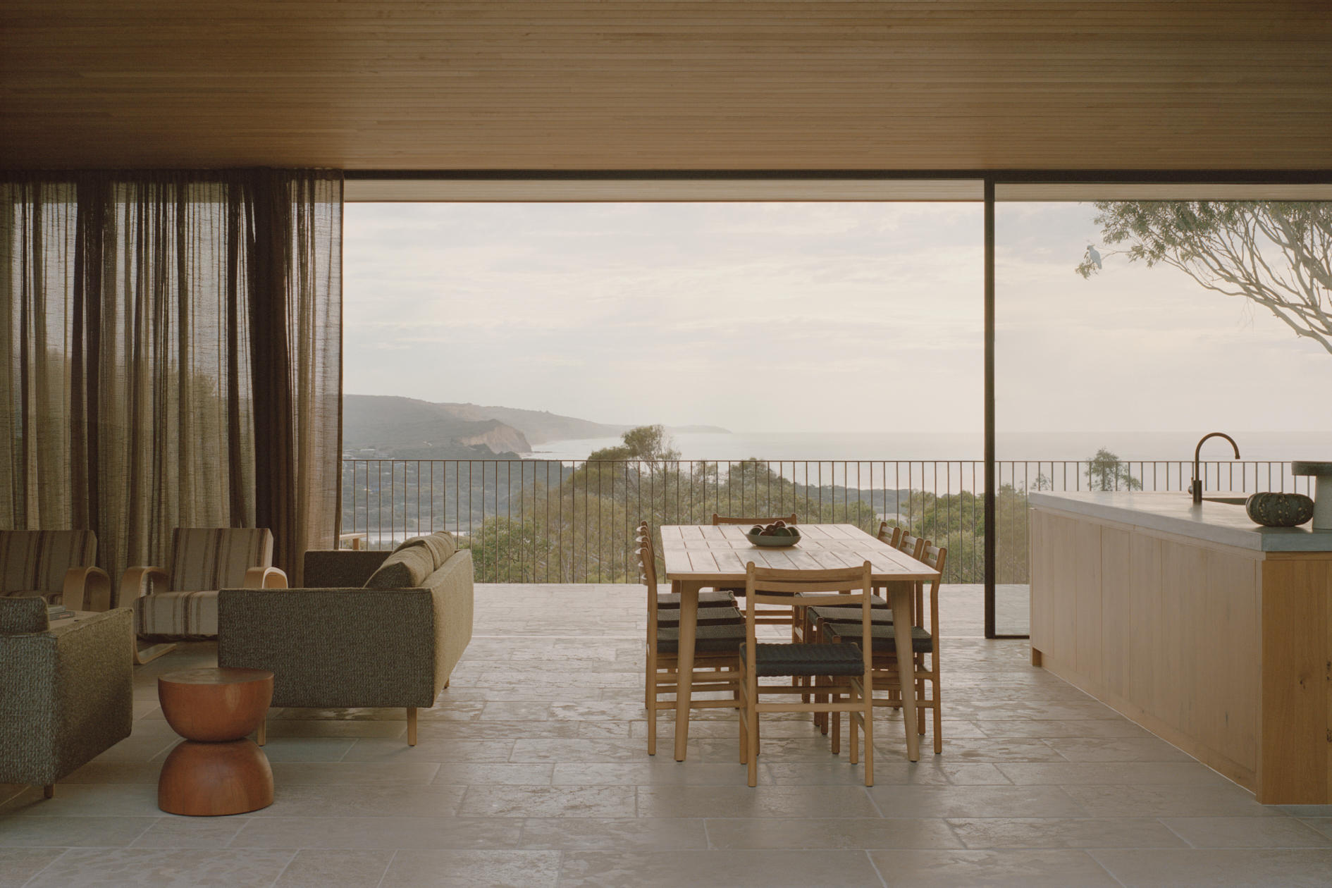 Open-plan living and dining space with full-height glazing opening to a coastal terrace overlooking distant hills