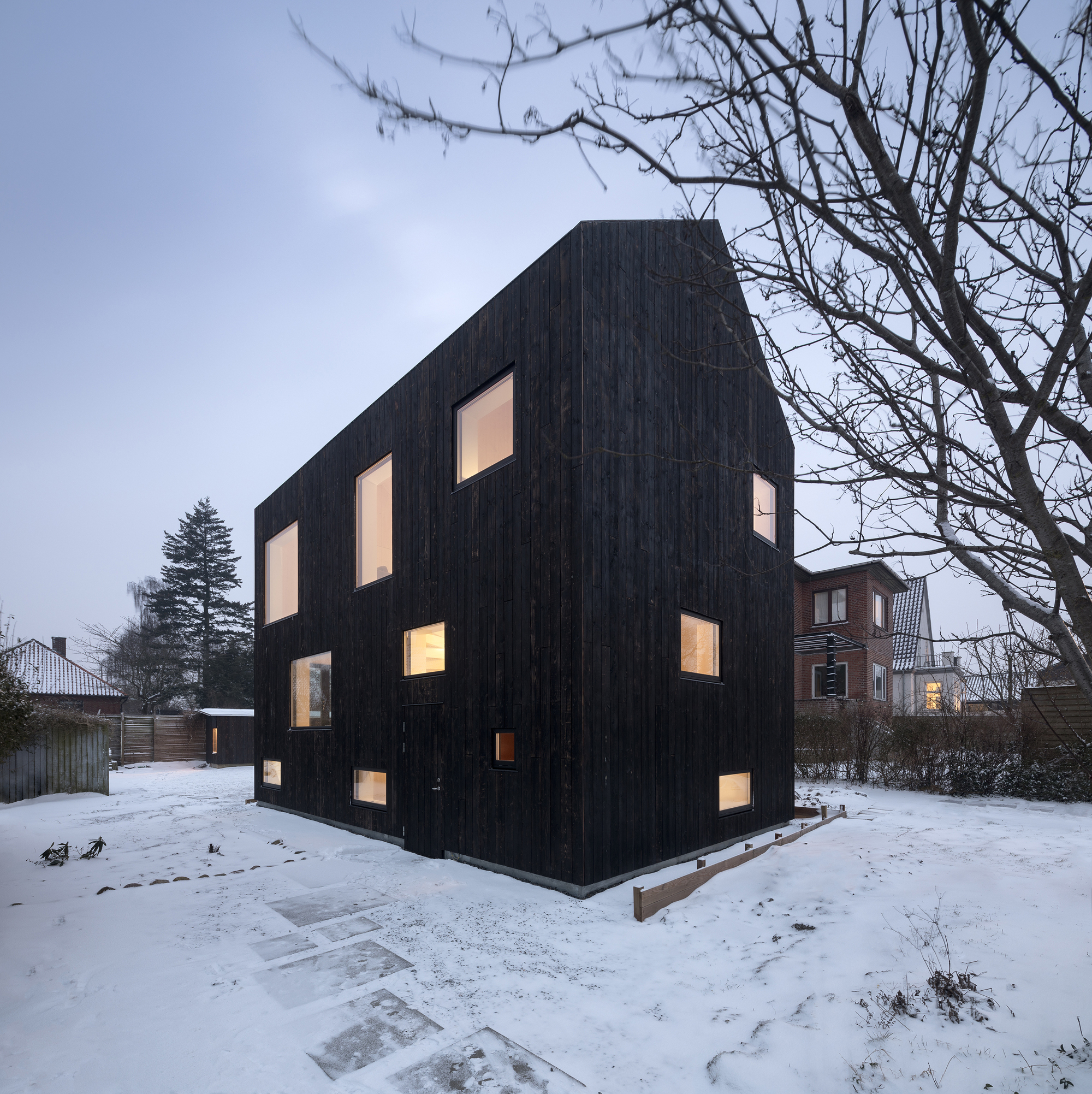 Corner view of the charred timber volume with illuminated windows in a snow-covered yard at twilight
