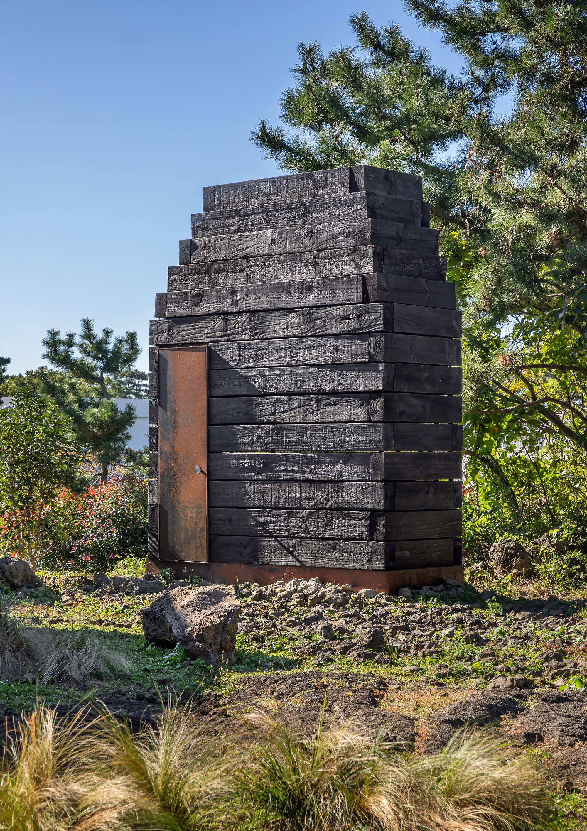 Charred timber facade with stepped profile and weathered steel doorway amid pine trees