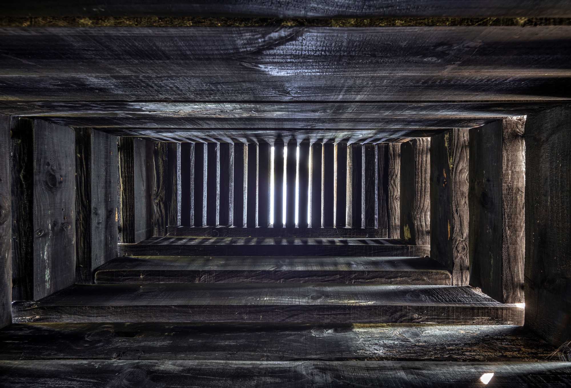 Looking upward through a timber stairwell with charred wood treads and vertical posts framing vertical light slots