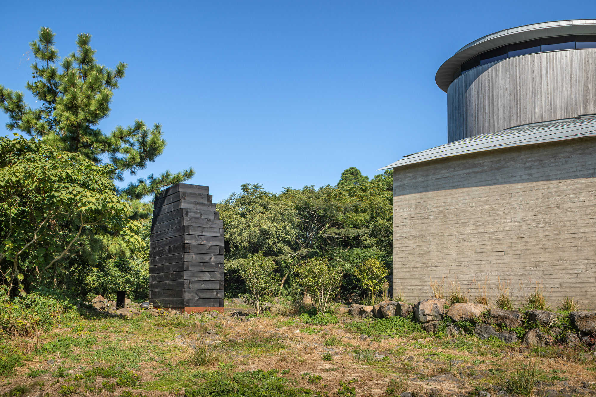 Exterior view of a stepped charred timber tower and cylindrical concrete volume surrounded by native grasses and trees
