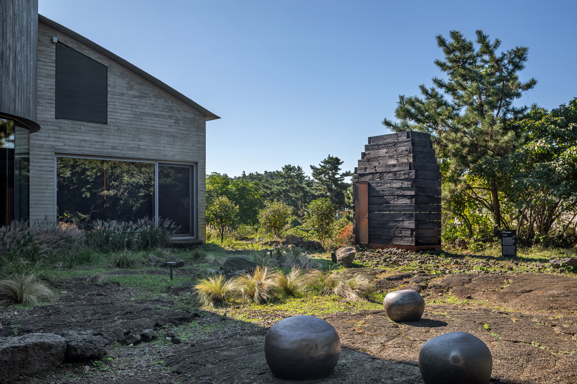 Garden courtyard with dark stone boulders, ornamental grasses, and charred timber tower beside timber-clad residence