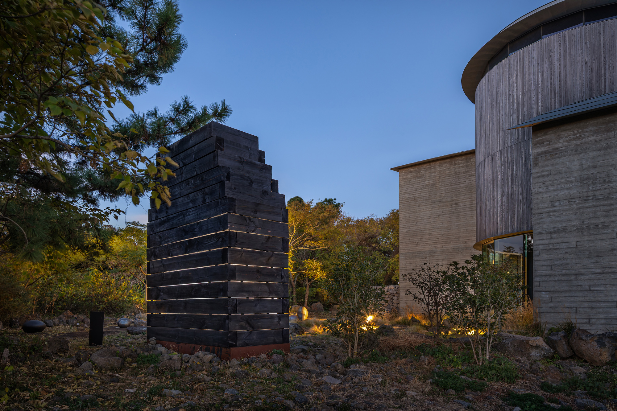 Charred timber tower with horizontal light strips at dusk beside cylindrical concrete volume and planted landscape