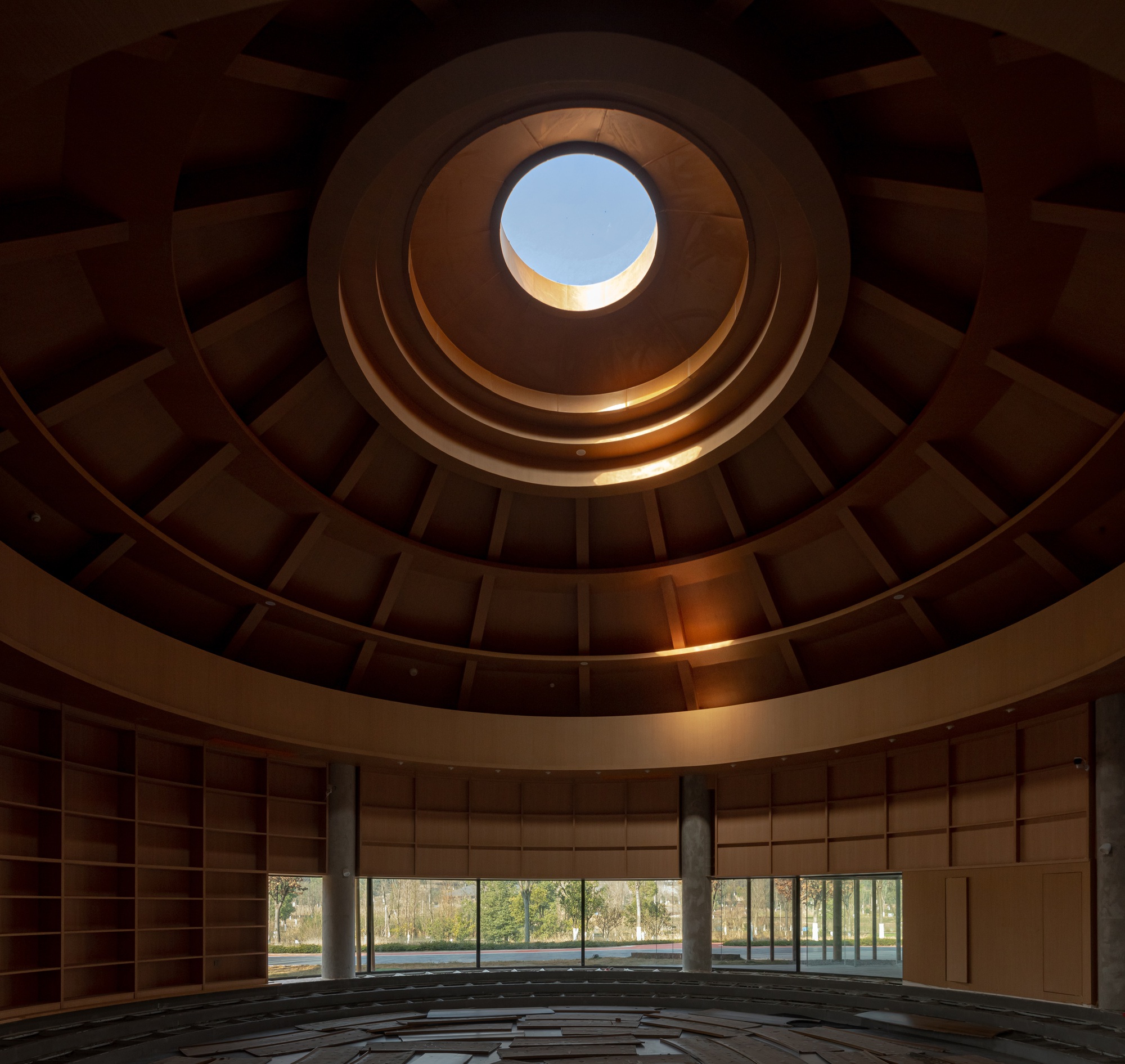 Looking up through a circular coffered dome with concentric timber rings and a central oculus framing blue sky