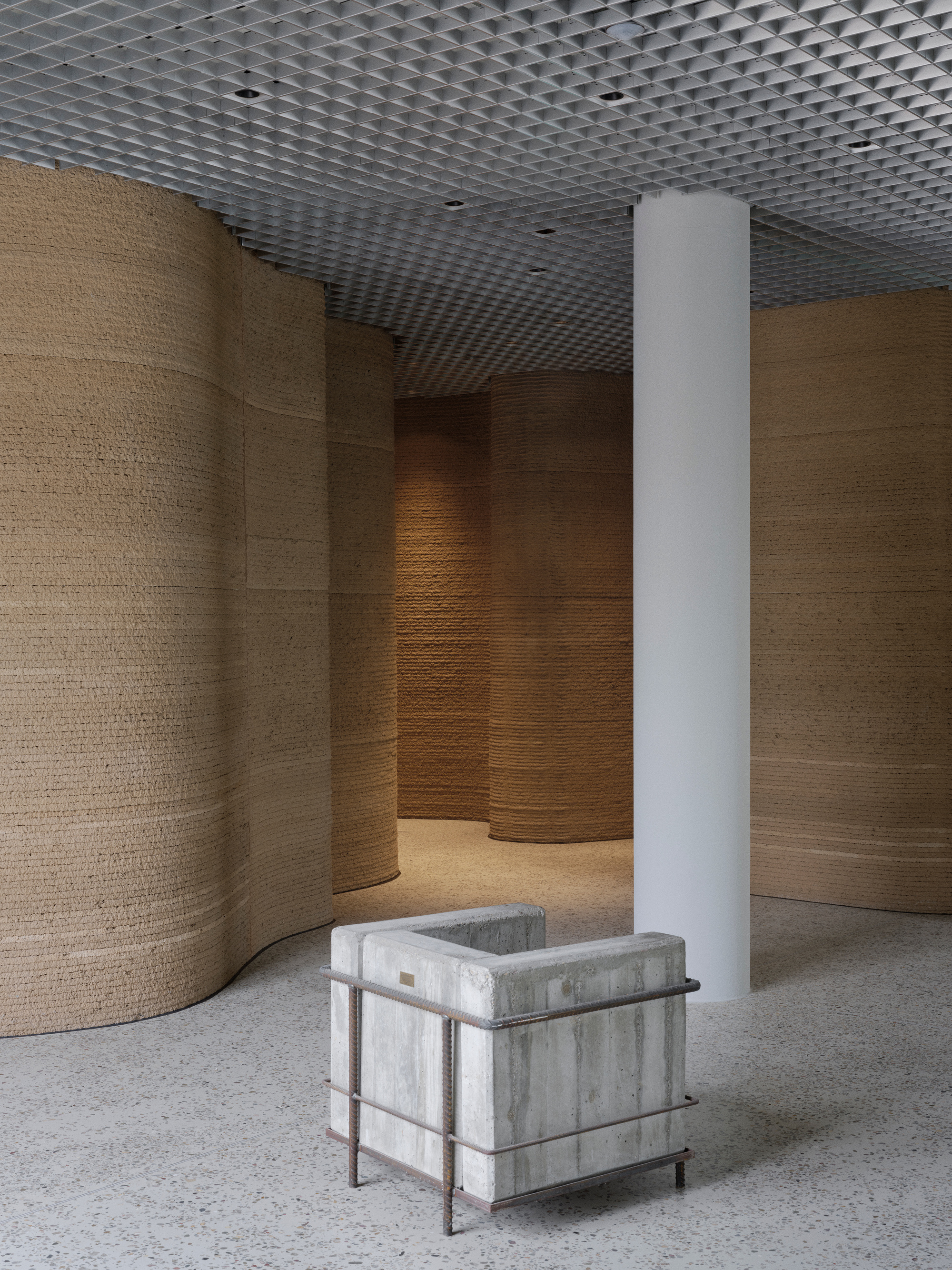 White cylindrical column beside concrete seating cube amid curved timber wall panels and open mesh ceiling