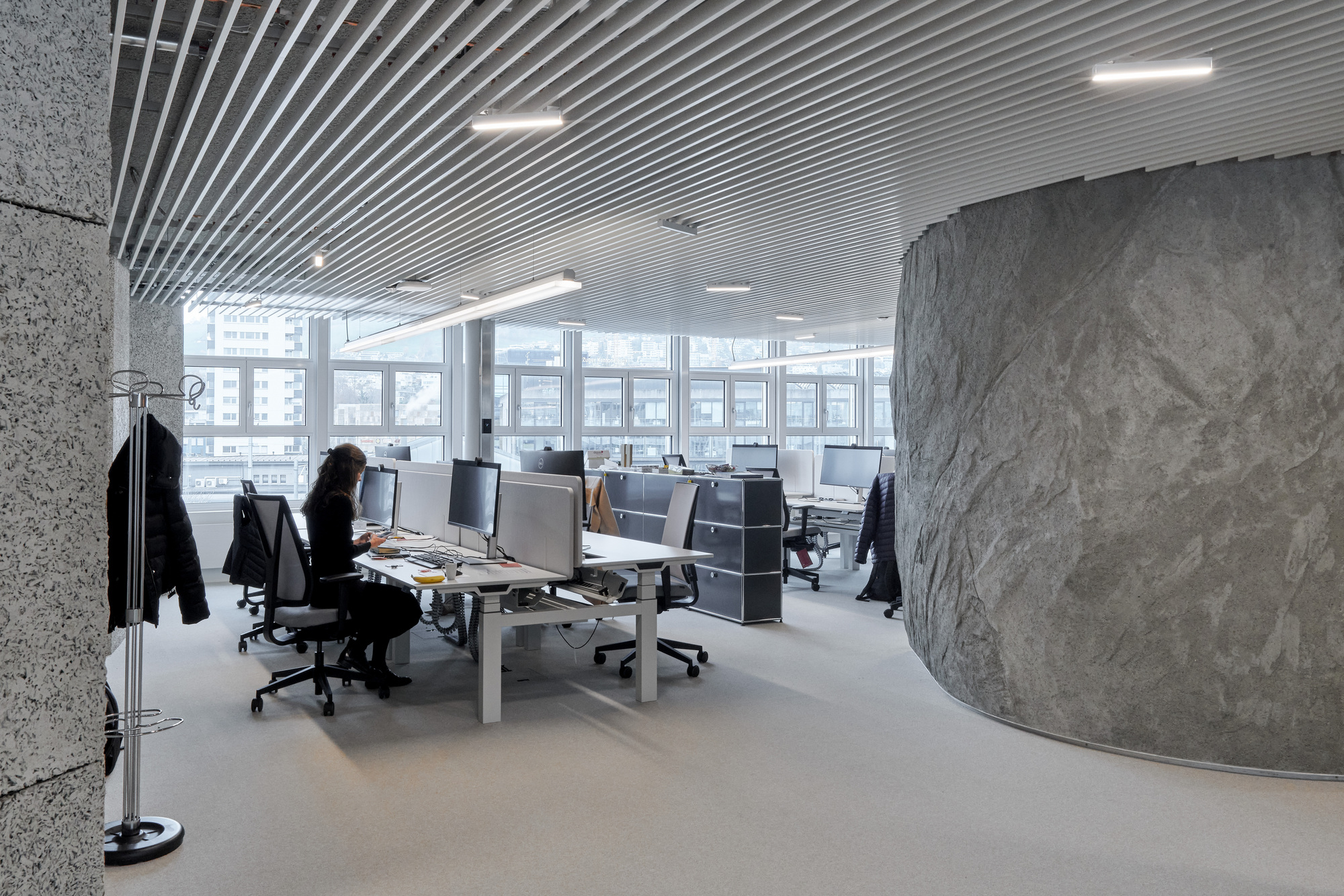 Open-plan office with workstations, curved grey stone wall, ribbed ceiling, and person seated at desk near windows