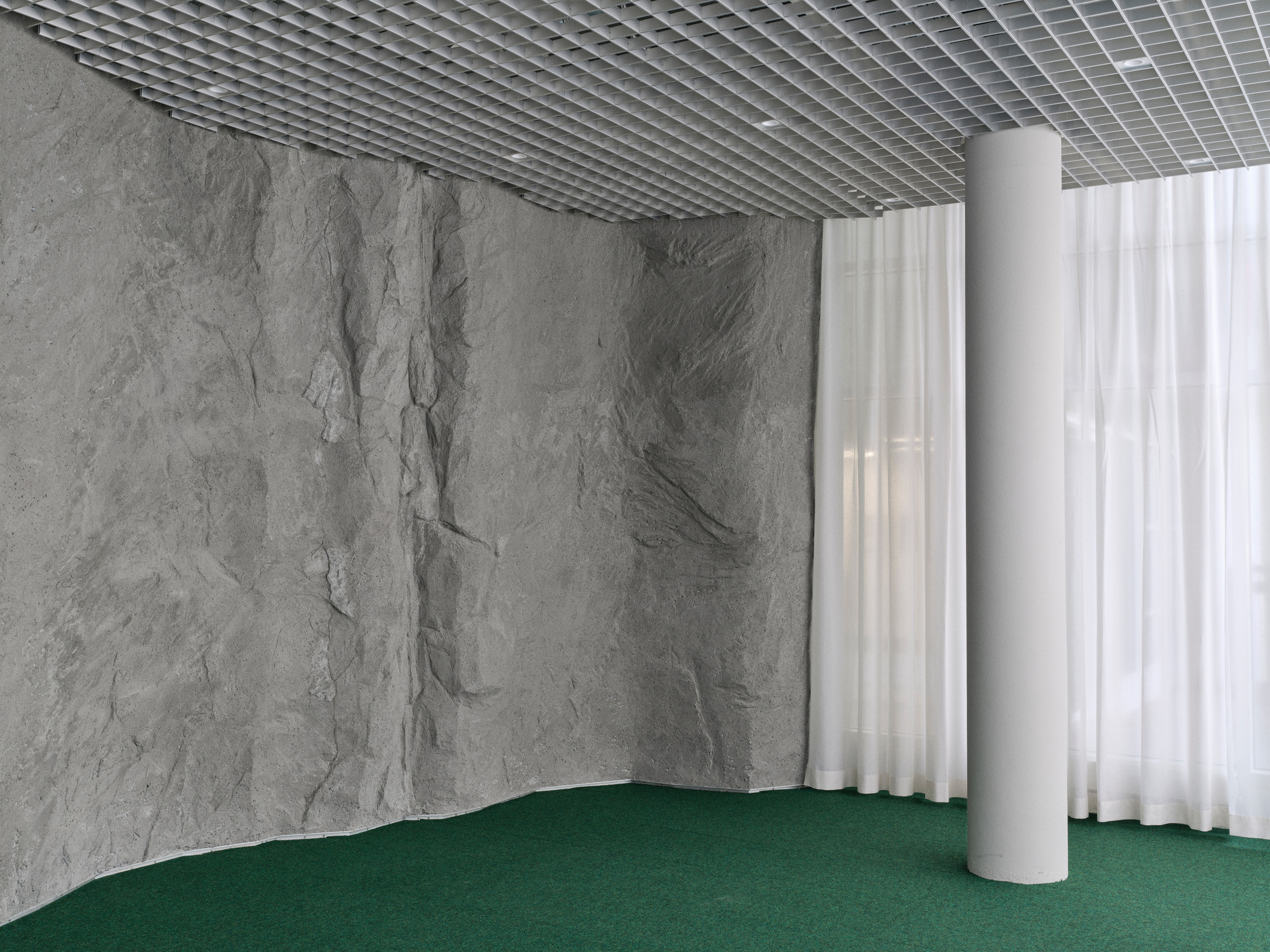 Corner with grey stone wall meeting white sheer curtain, green carpet flooring, and open grid metal ceiling