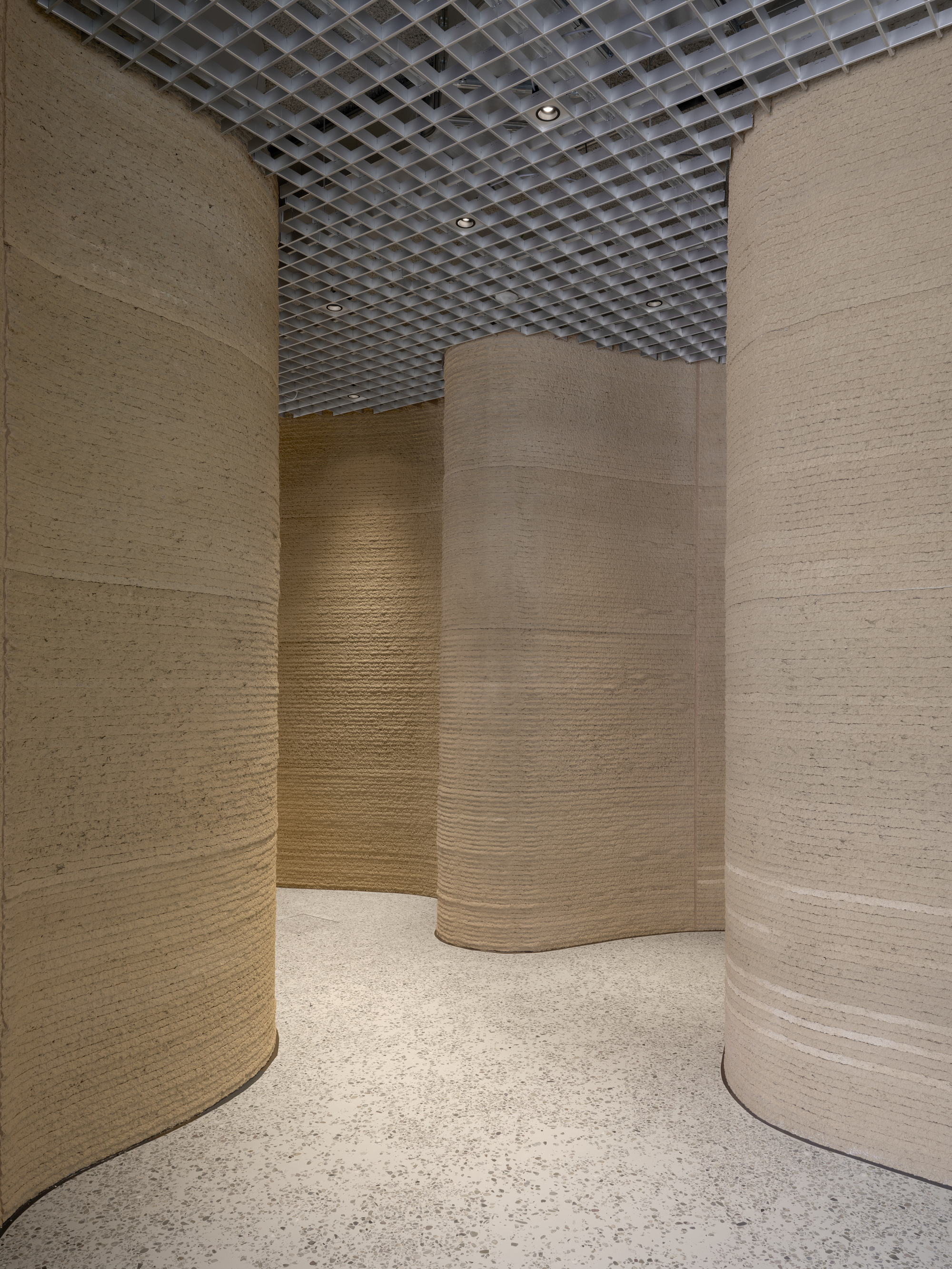 Passage between three curved copper-paneled columns with terrazzo flooring and open grid ceiling above