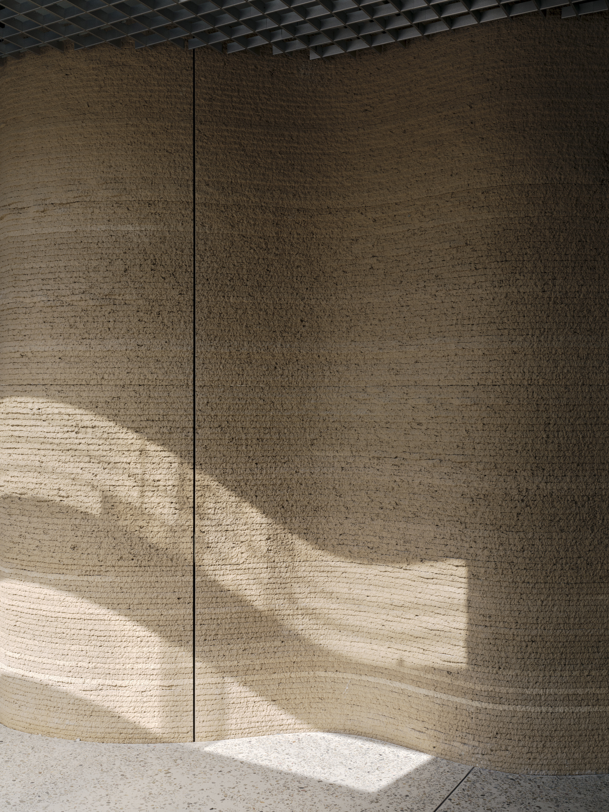 Curved rammed earth wall panels catching diagonal sunlight beneath perforated metal ceiling grid