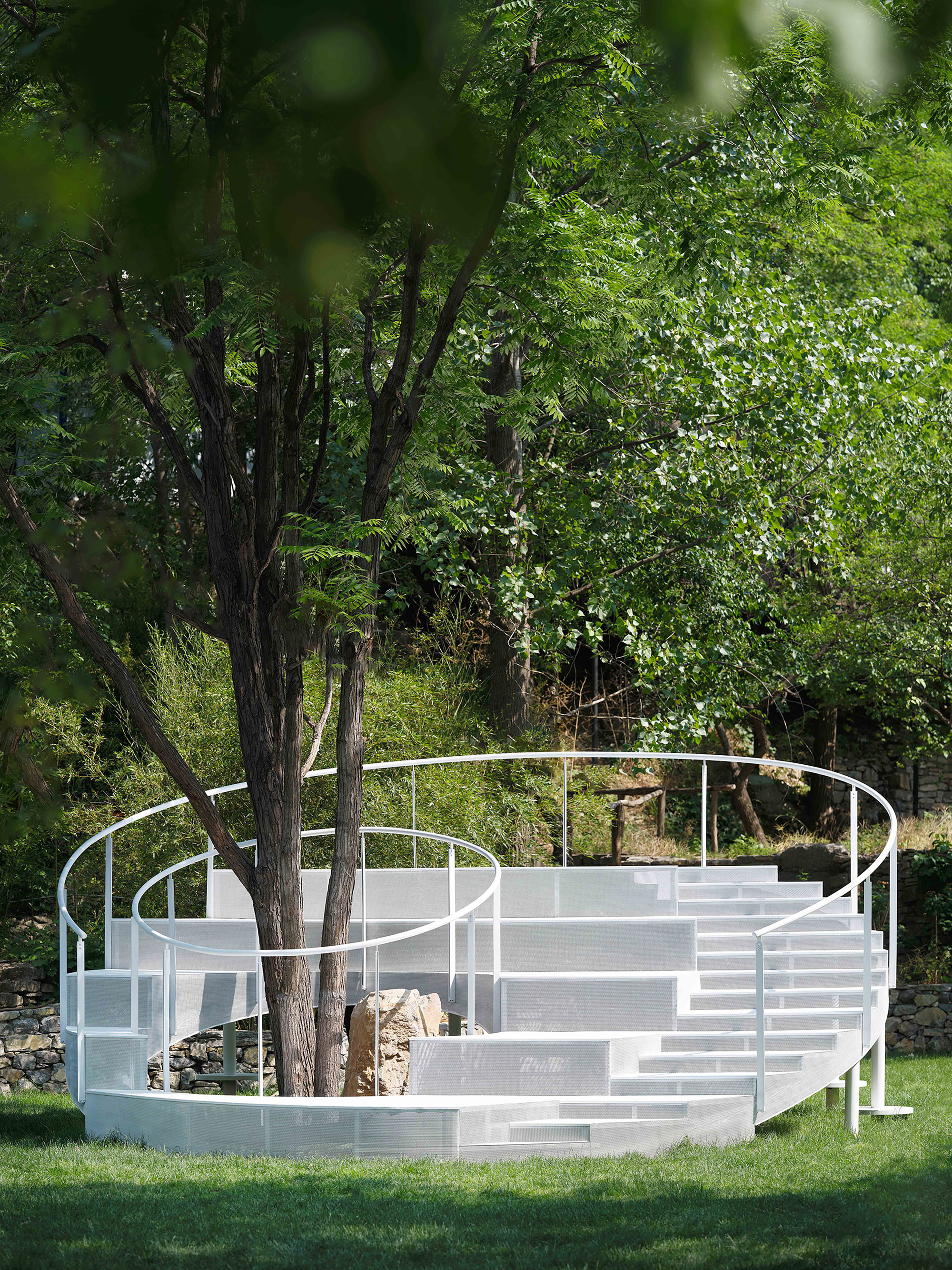 White steel amphitheater structure with tiered seating wrapped around existing trees in a summer lawn