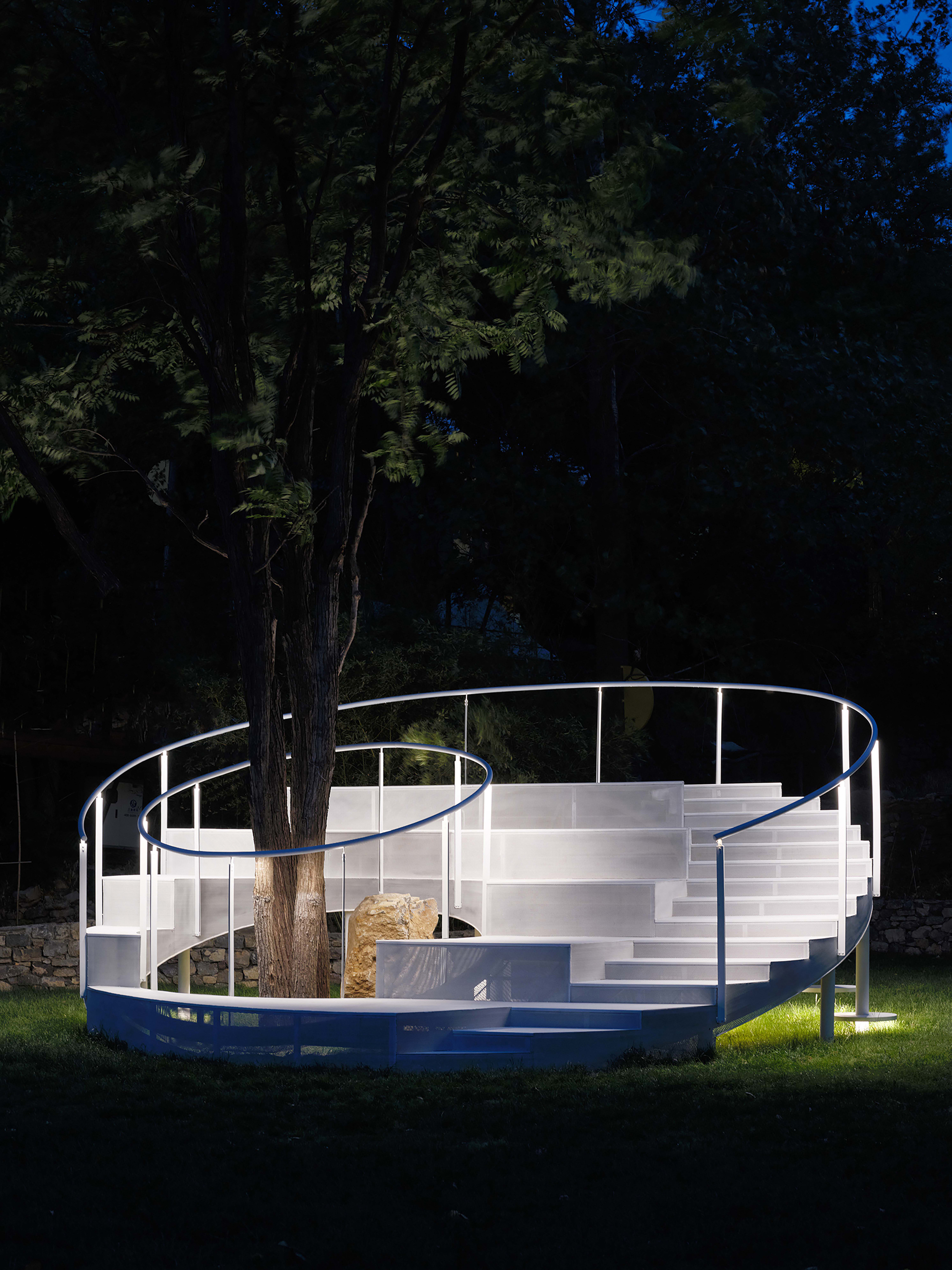Illuminated spiral staircase structure encircling a tree at night with rock at center
