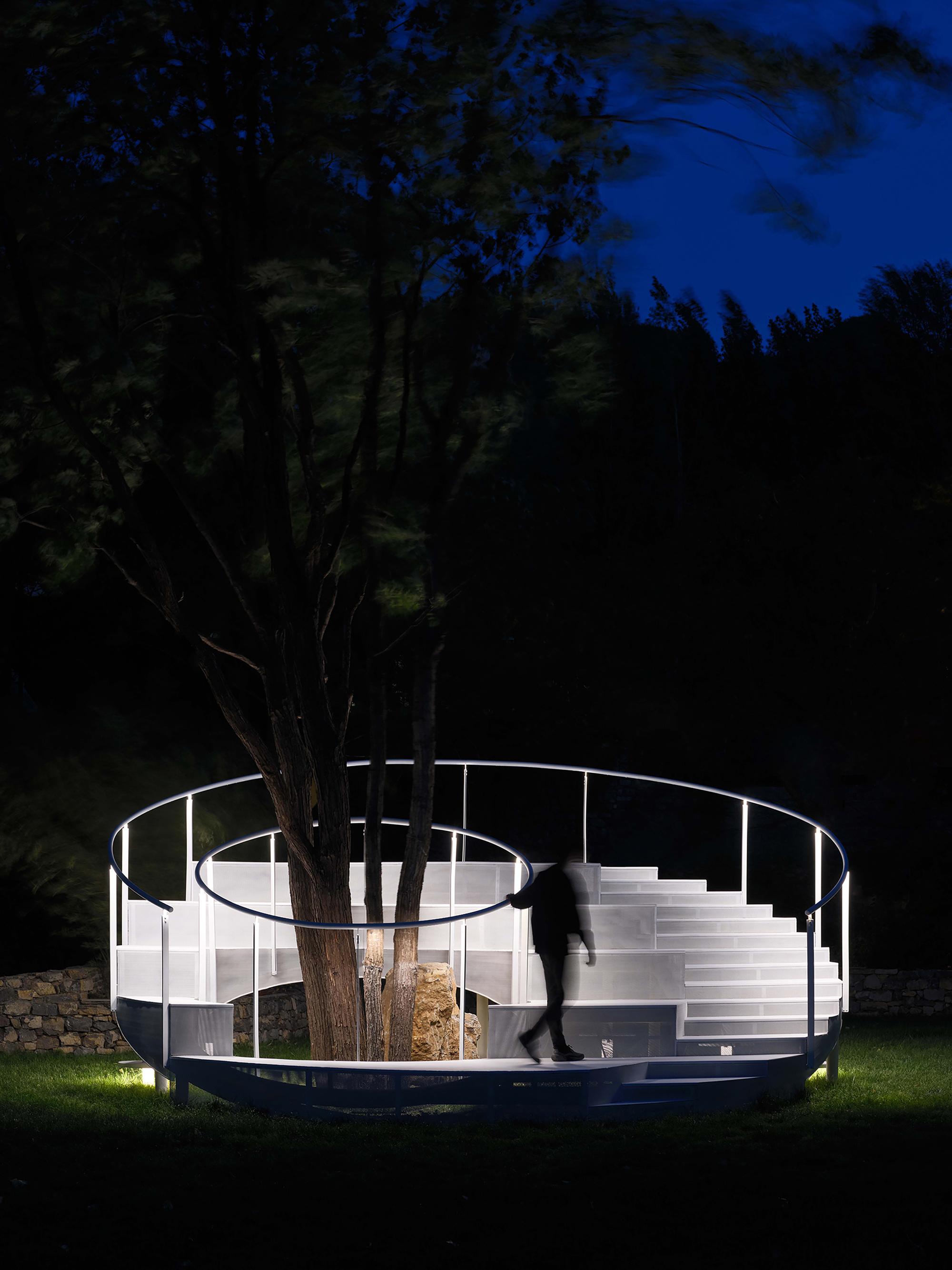 Illuminated circular pavilion with white metal railings and stairs wrapping around a tree at dusk