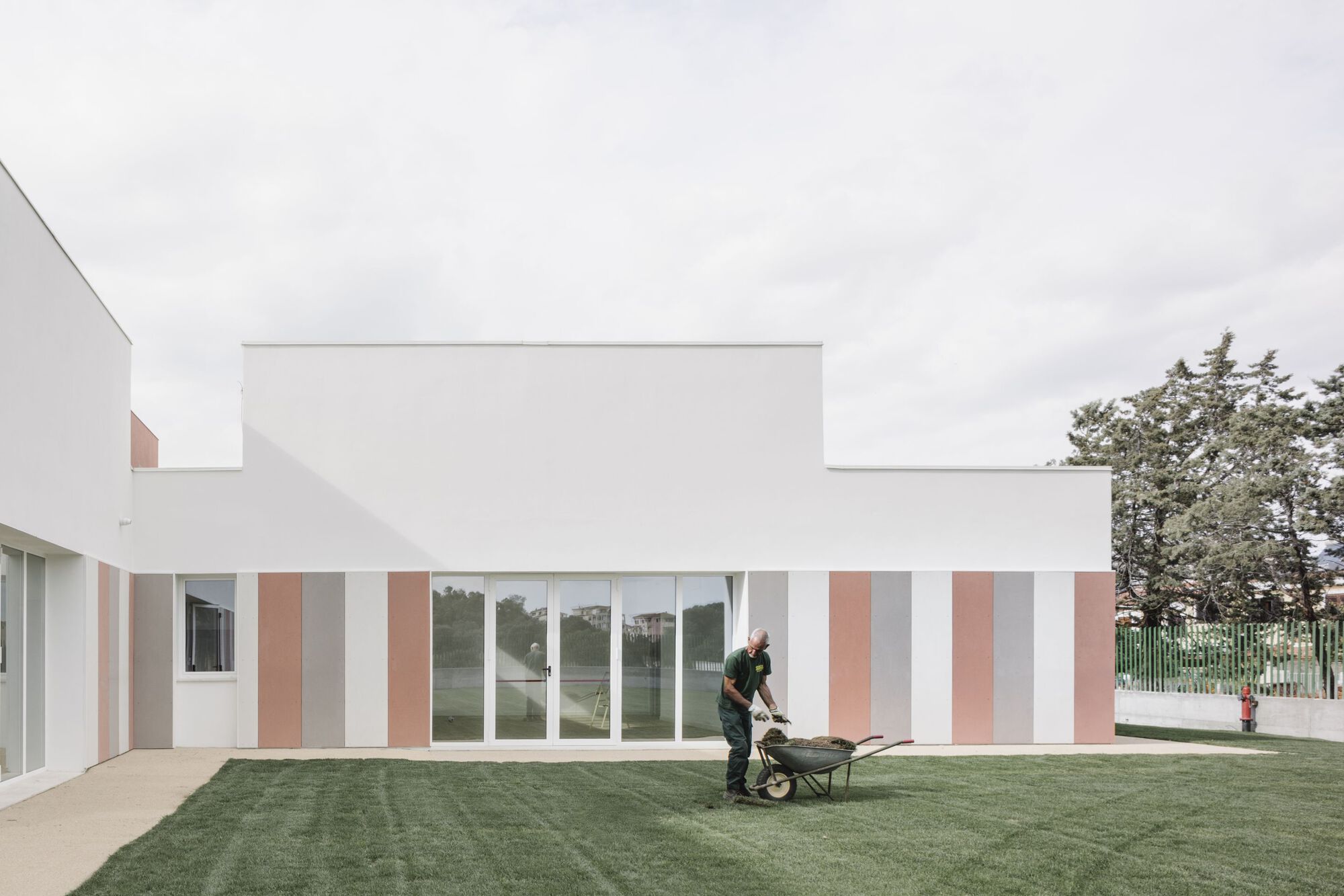 Vertically striped facade in white, grey and terracotta panels with a worker pushing a wheelbarrow on the lawn