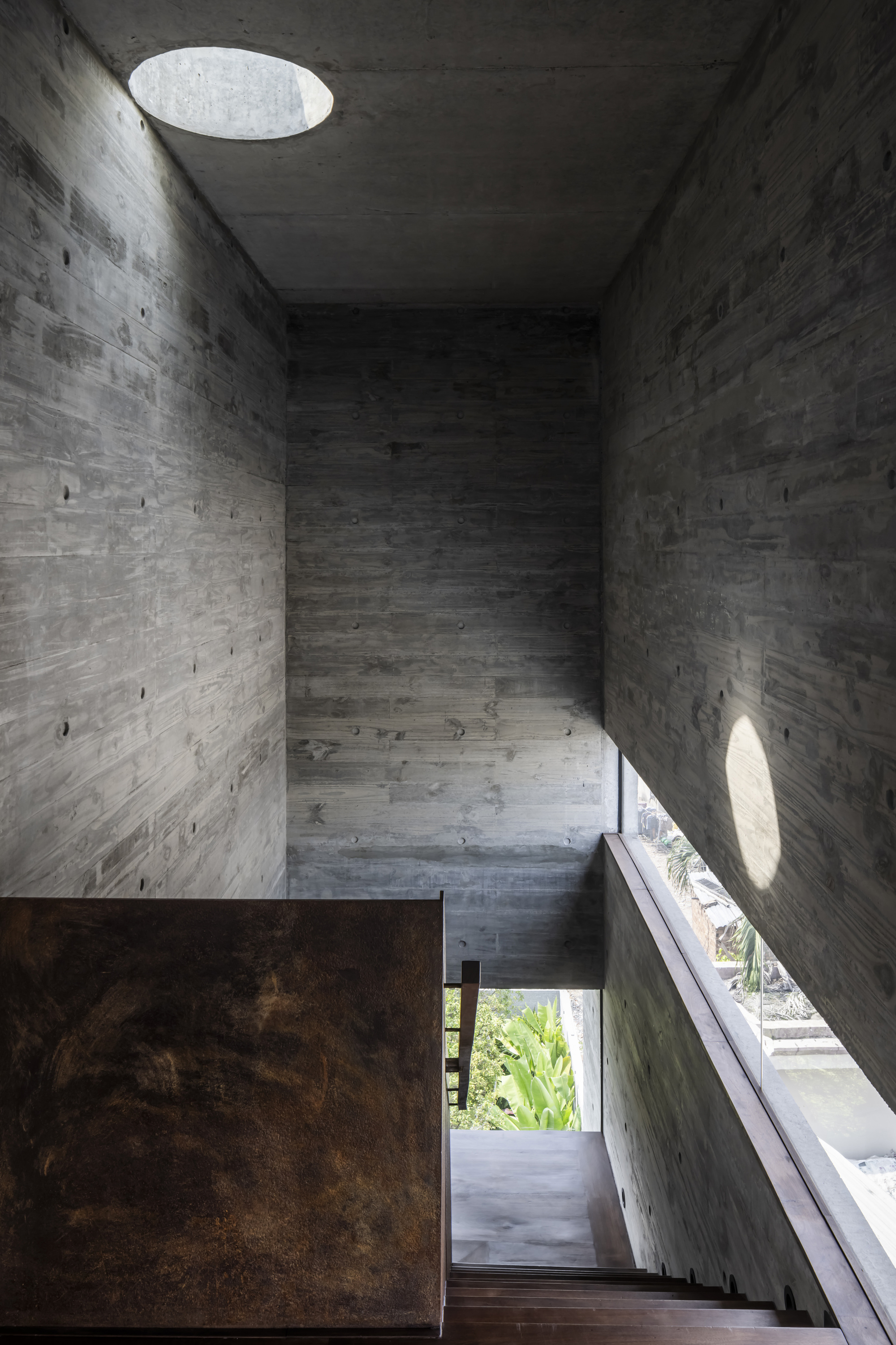 Board-formed concrete stairwell with circular ceiling oculus and angled shadows from afternoon sun