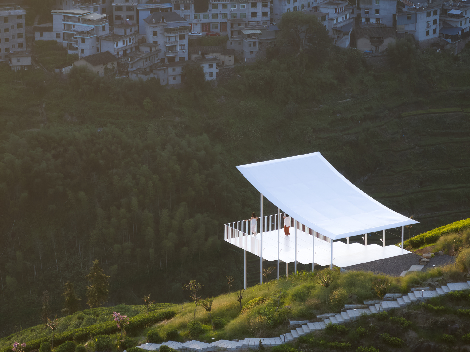Elevated pavilion with curved white roof and slatted railing set among forested hillside with distant buildings