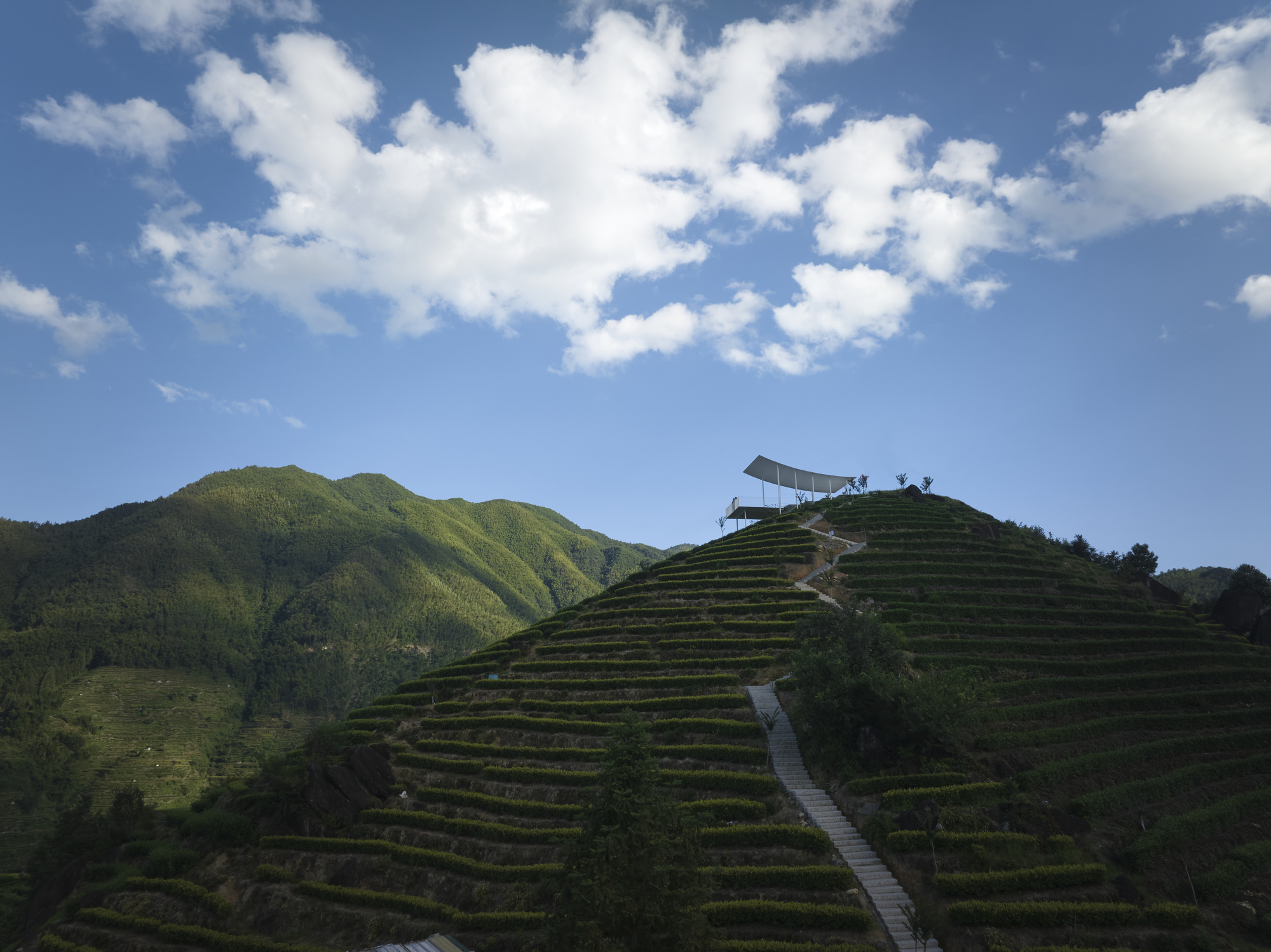 White canopy pavilion perched atop terraced hillside under blue sky with cumulus clouds and mountain backdrop