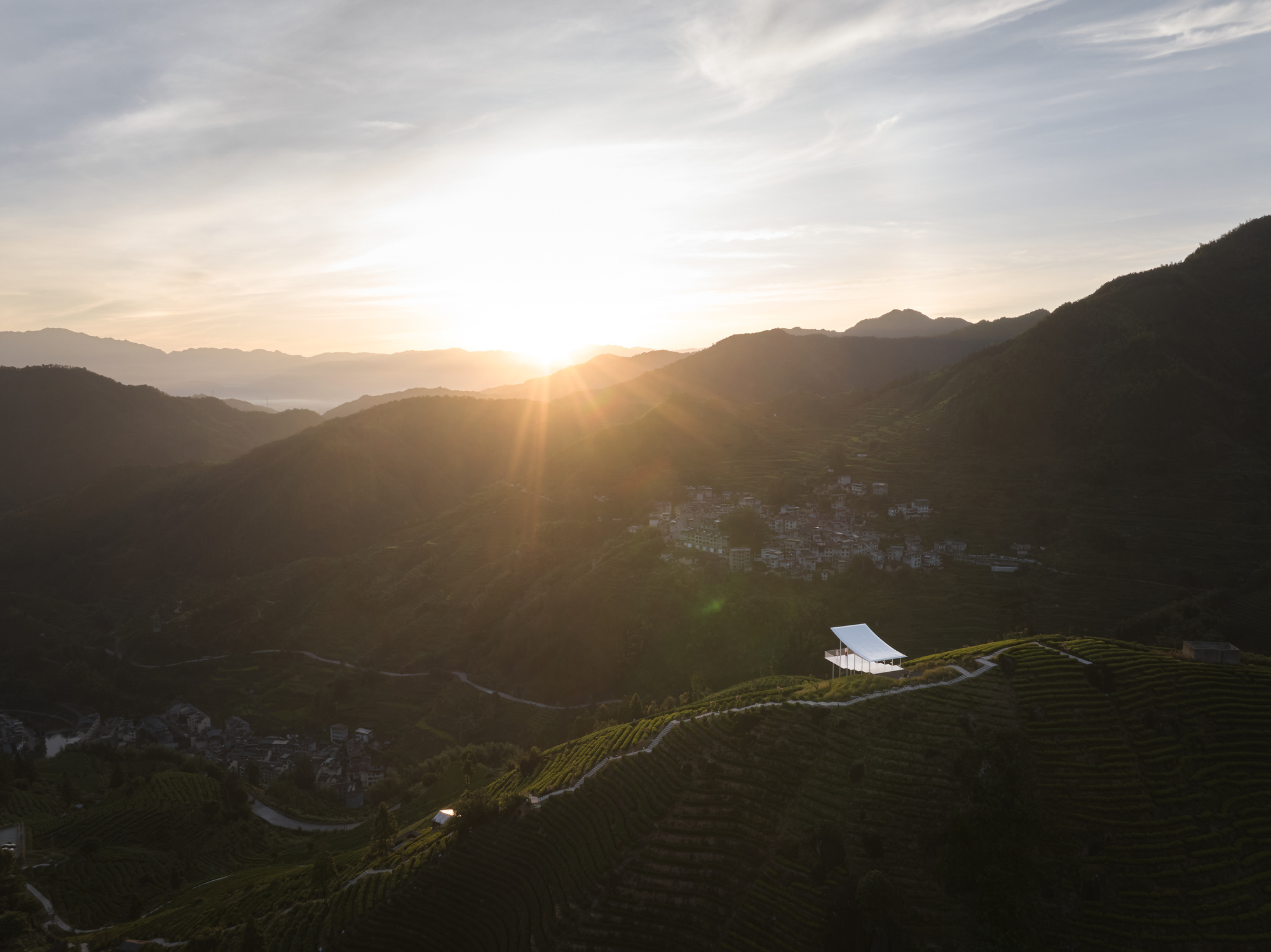 White pavilion on hillside plantation at sunset with distant mountain village in shadow