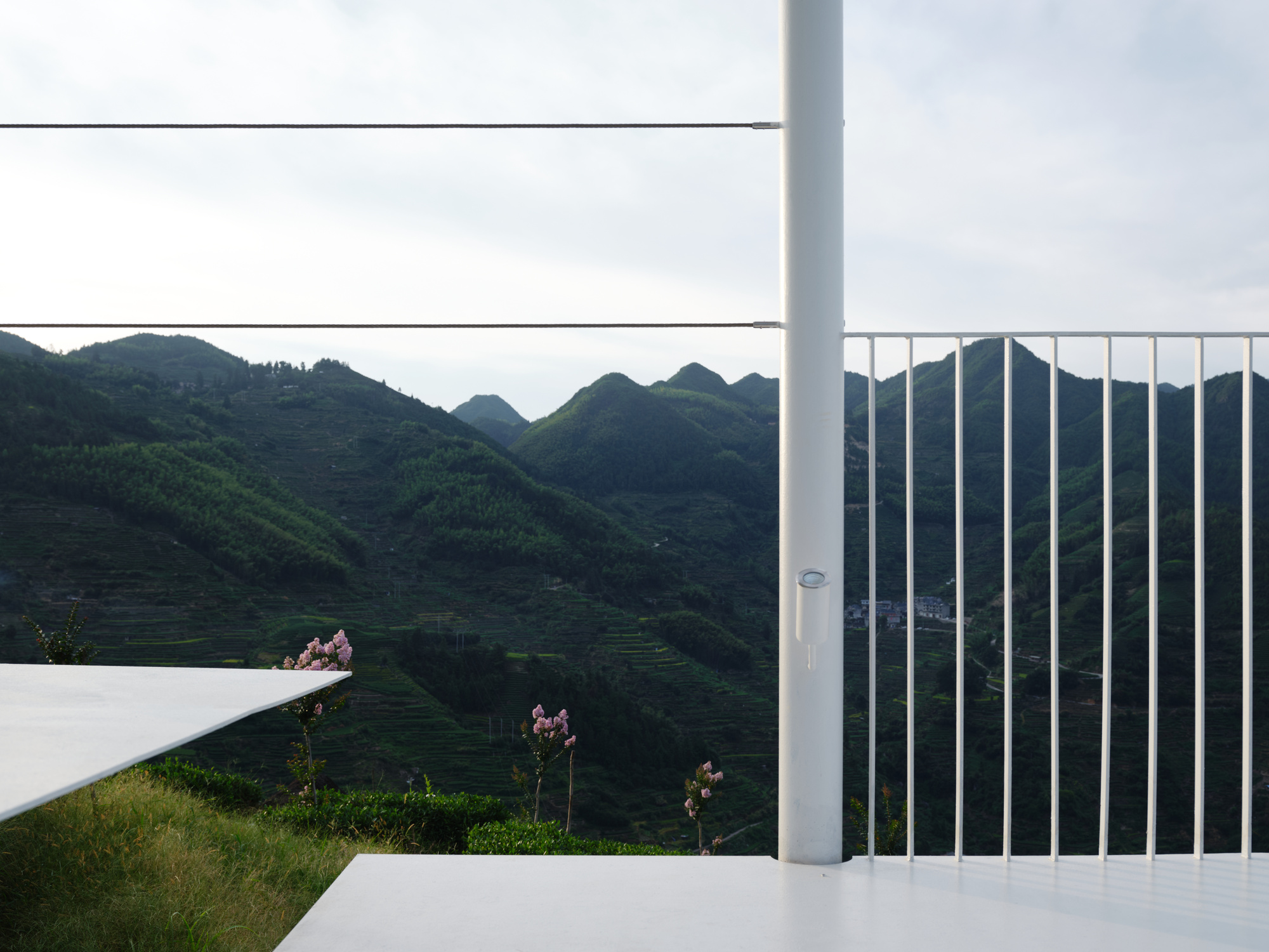 Vertical white railing and horizontal cable rails framing a view of green mountain slopes at dusk
