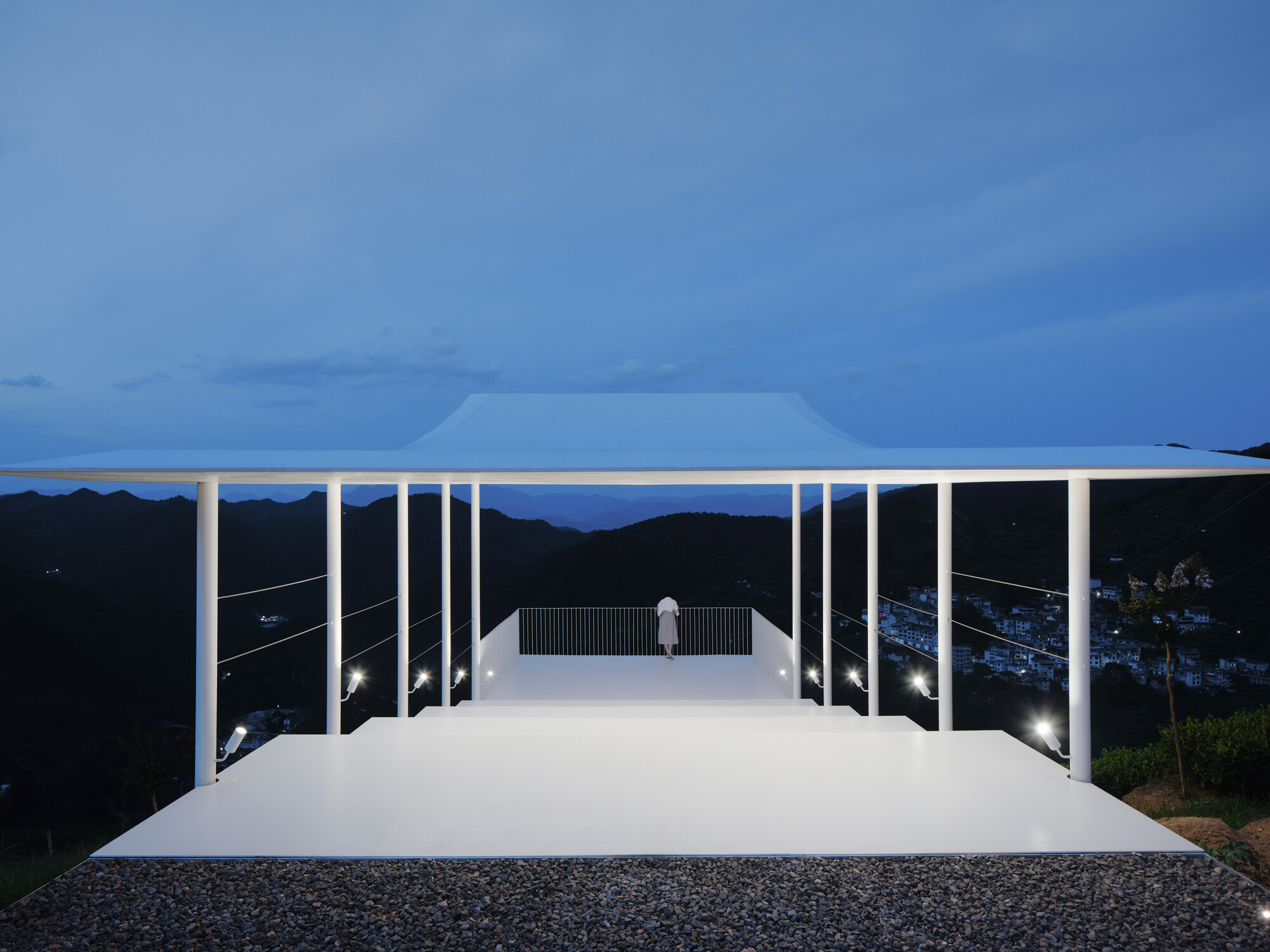 Elevated white pavilion with slender columns and a cantilevered roof at dusk overlooking mountains