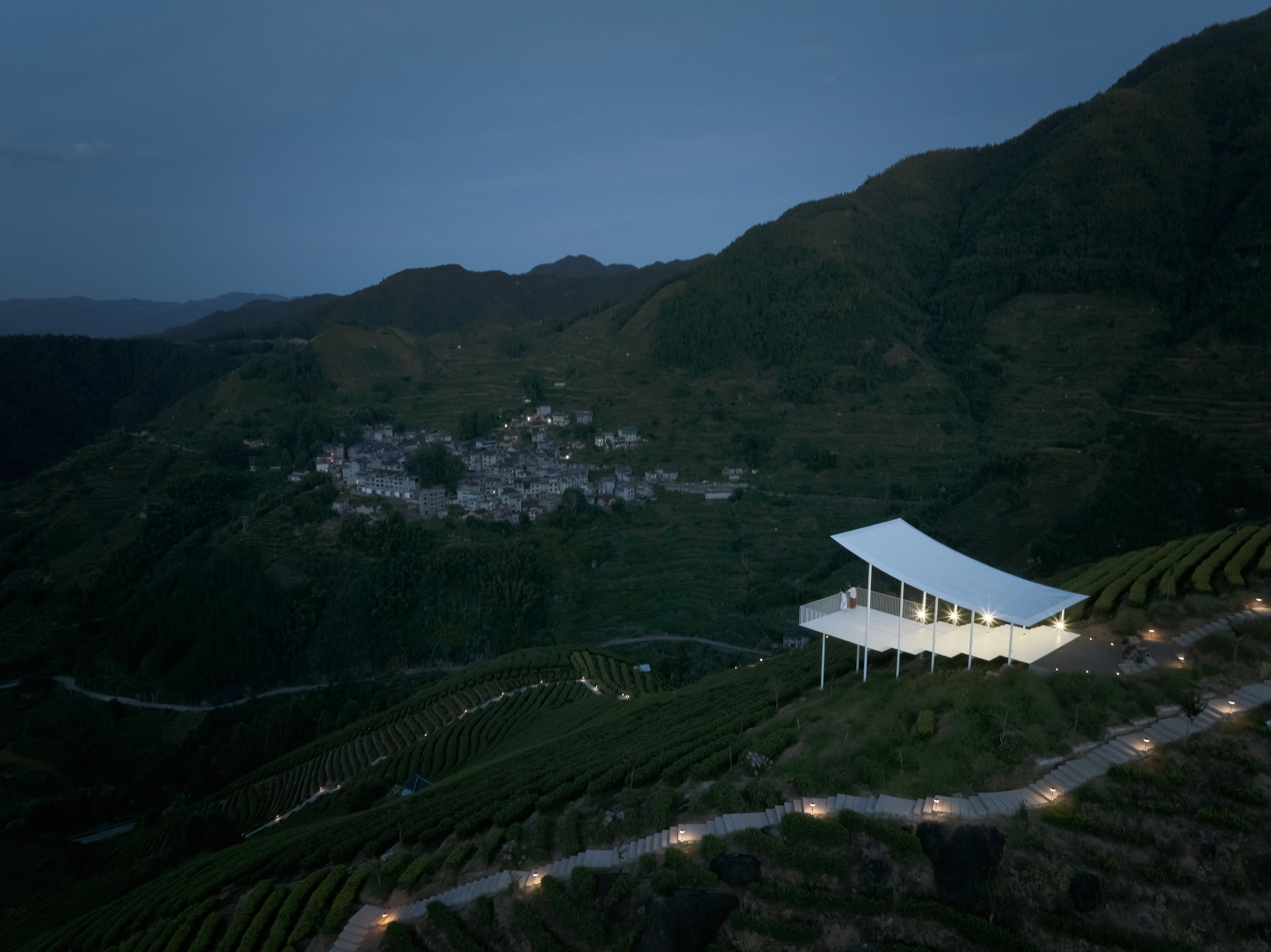 Aerial view of the pavilion perched on terraced tea fields above a hillside village at twilight
