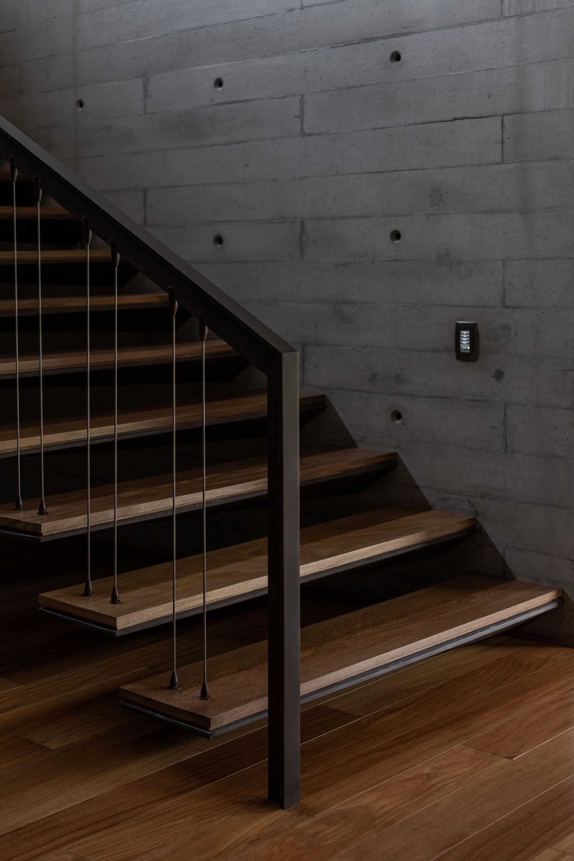 Timber staircase with steel cable railing against a board-formed concrete wall in low light