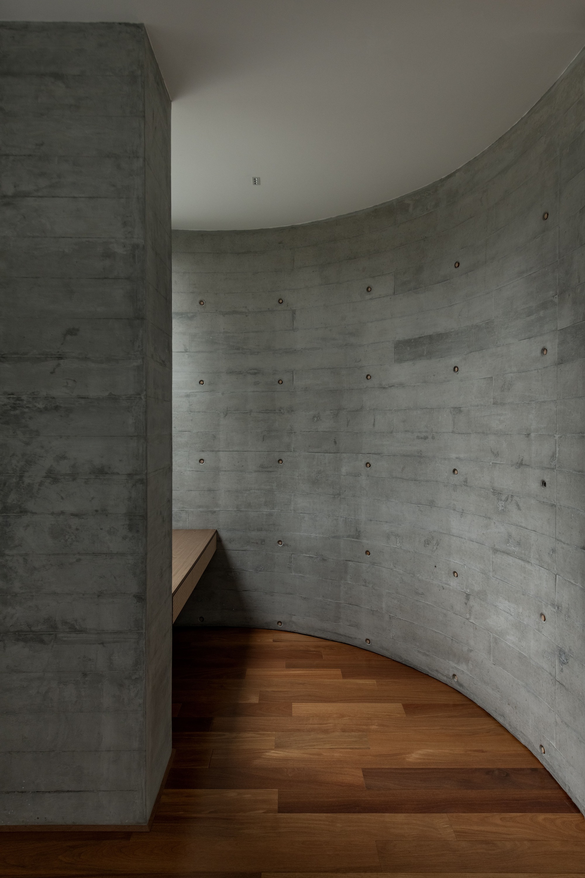 Curved board-formed concrete wall with timber bench and wooden flooring wrapping the interior space