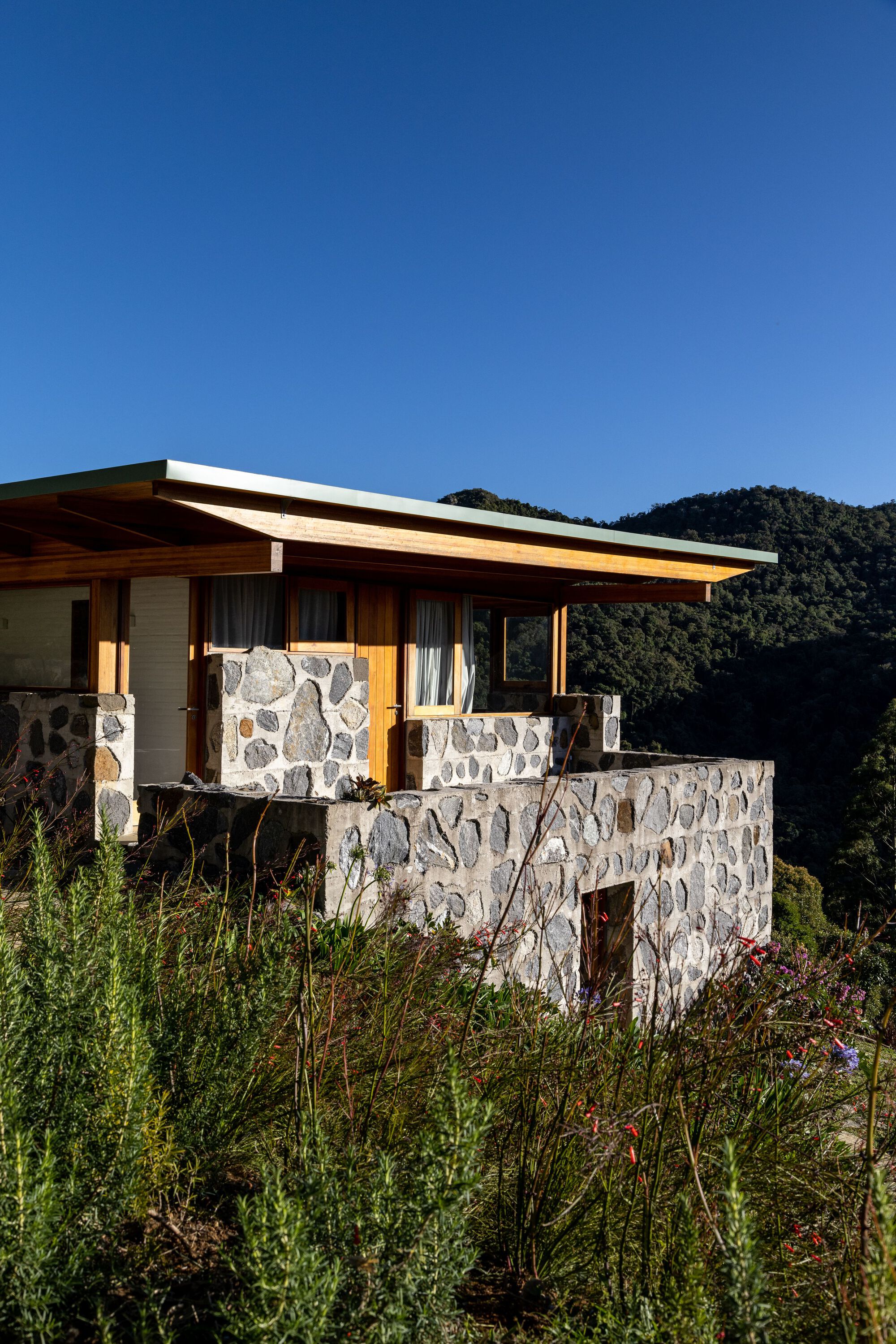 Two-level facade with river stone walls and timber framing surrounded by wildflowers under clear blue sky