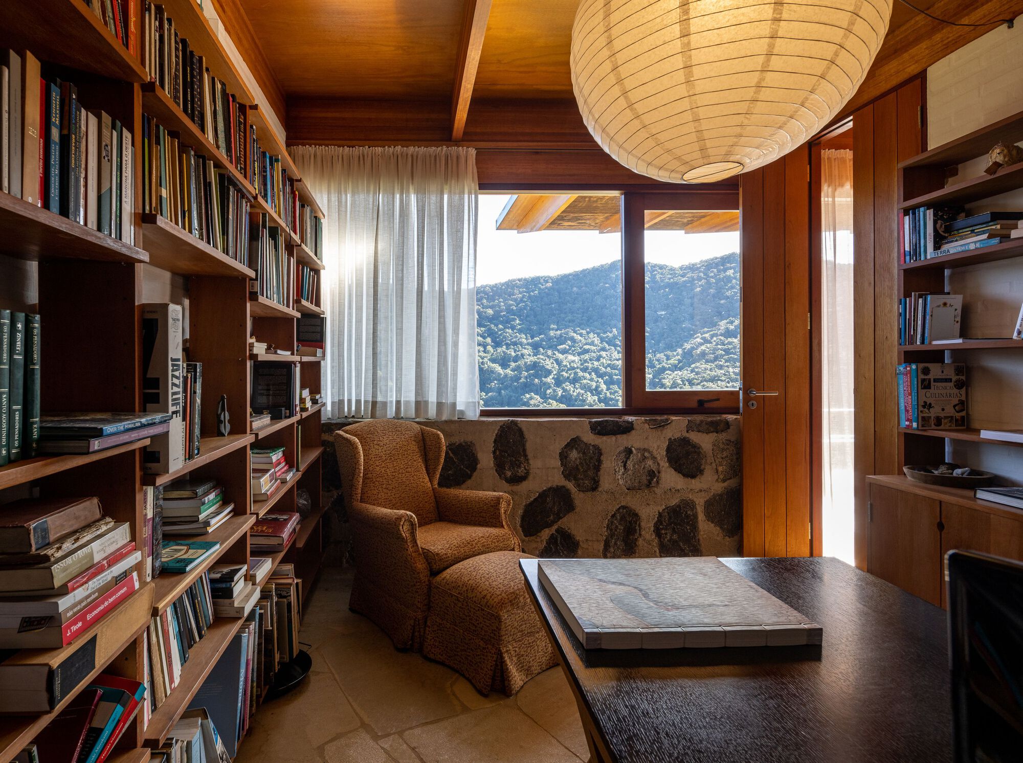 Library with built-in timber bookshelves and glazed doors opening to hillside views beyond the fieldstone wall