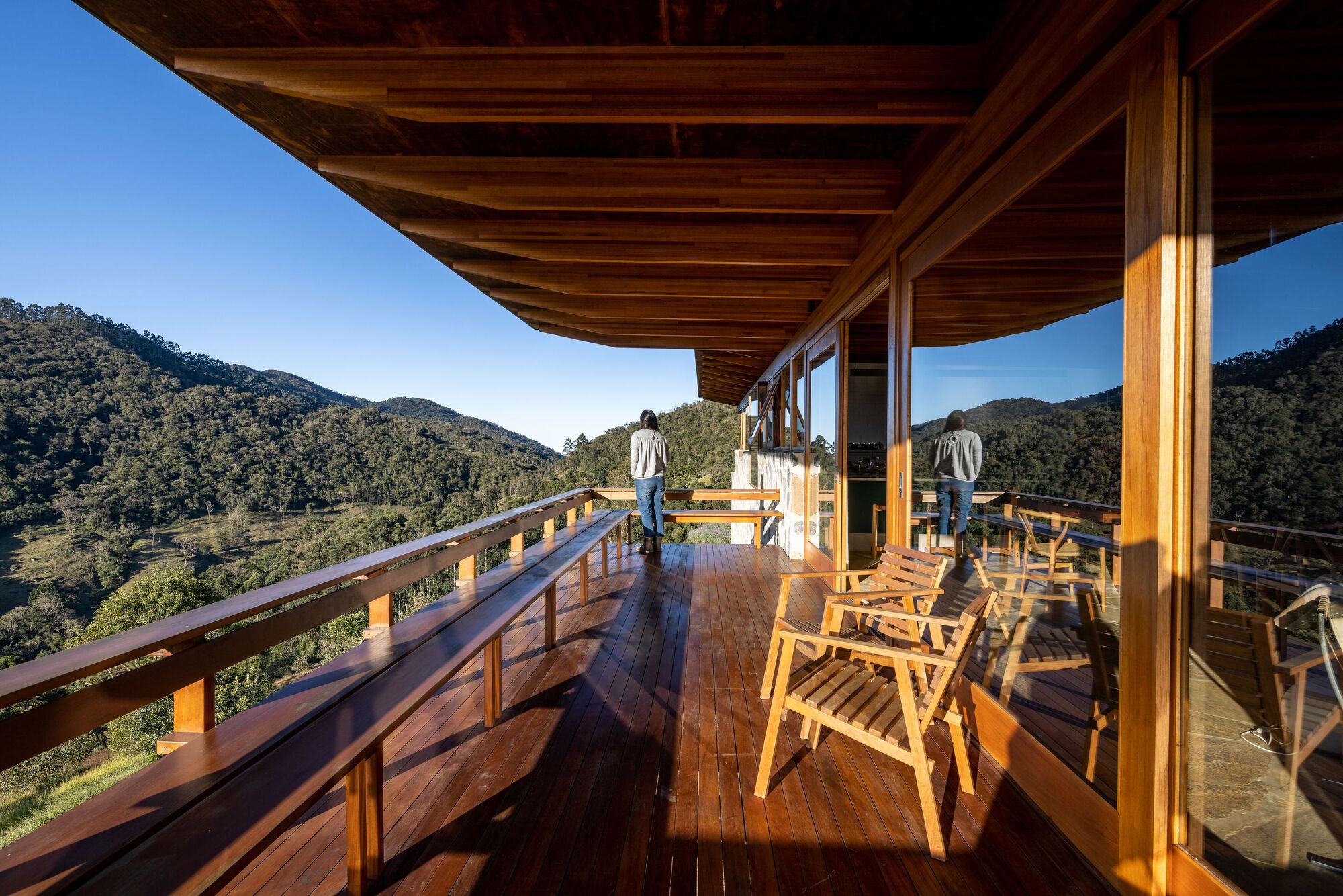 Covered deck with exposed timber rafters and railing where two figures look out over forested valley
