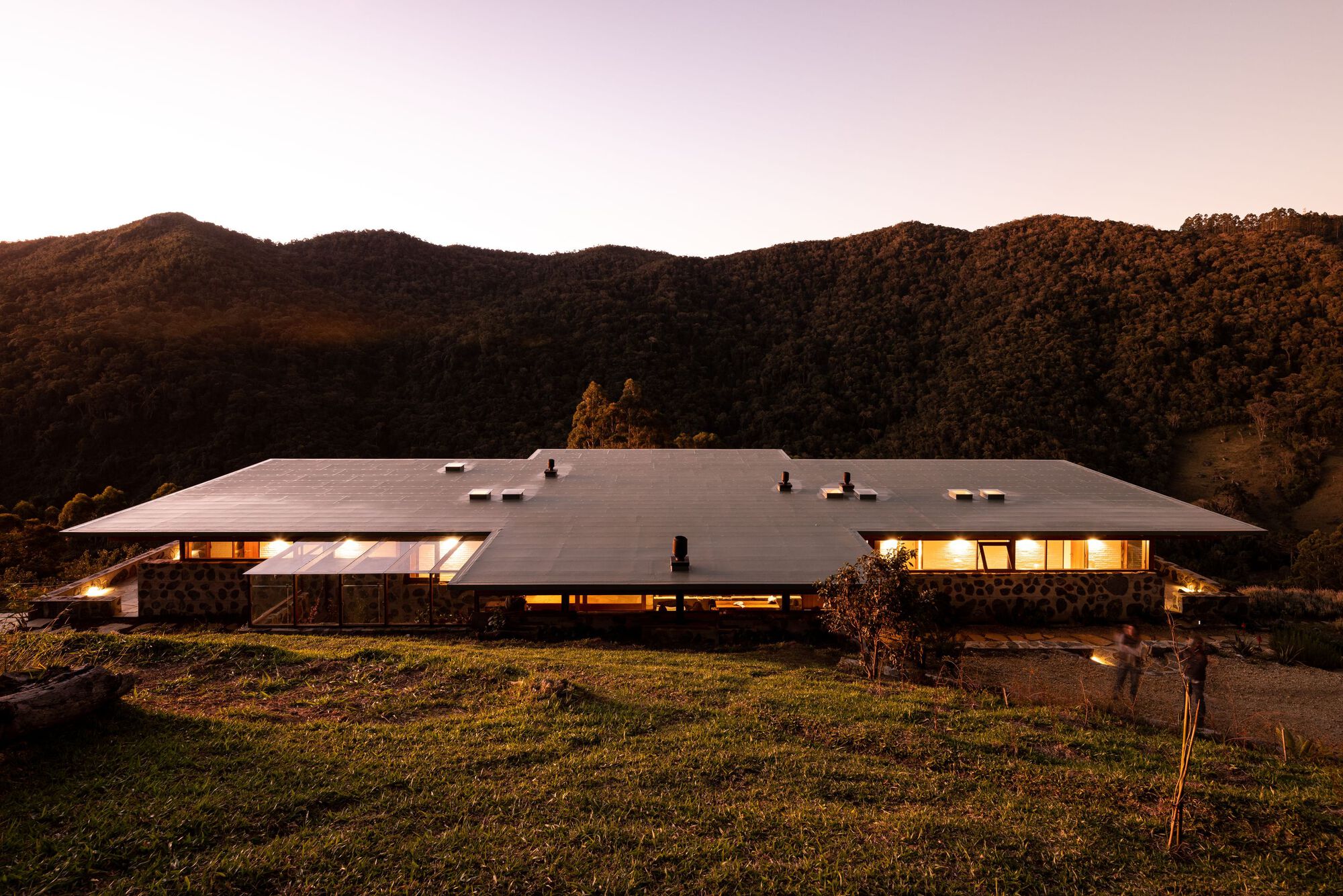 Low-slung residence with broad overhanging roof set against forested hillside at dusk
