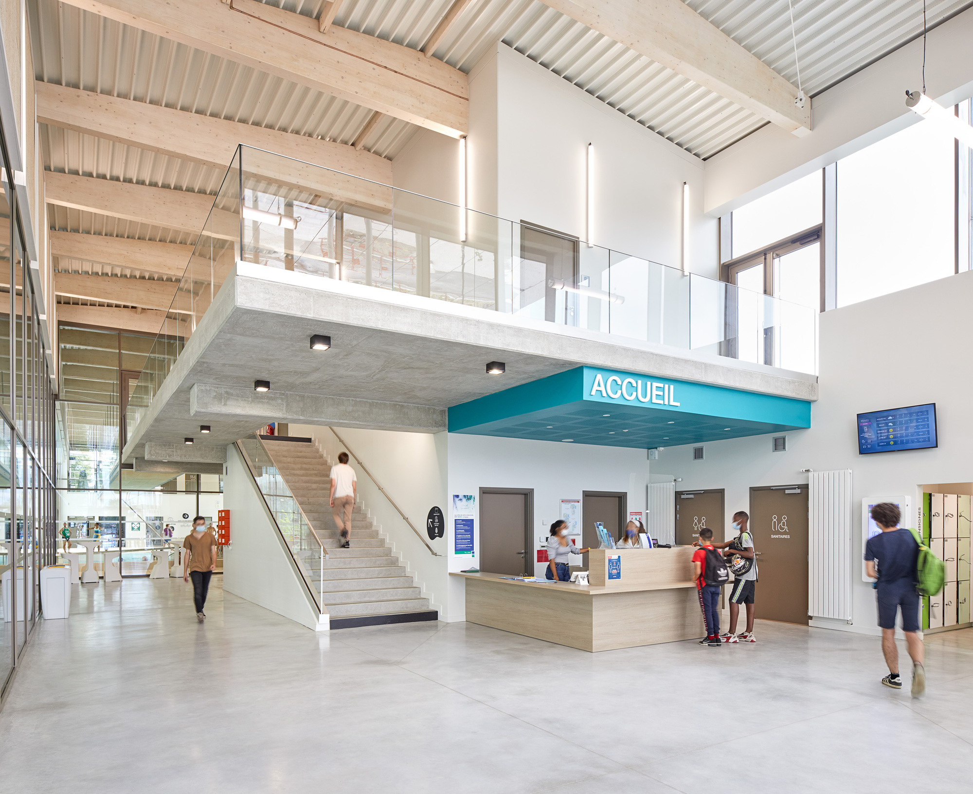 Double-height reception lobby with timber-slatted ceiling, mezzanine walkway, and visitors at the wooden desk below