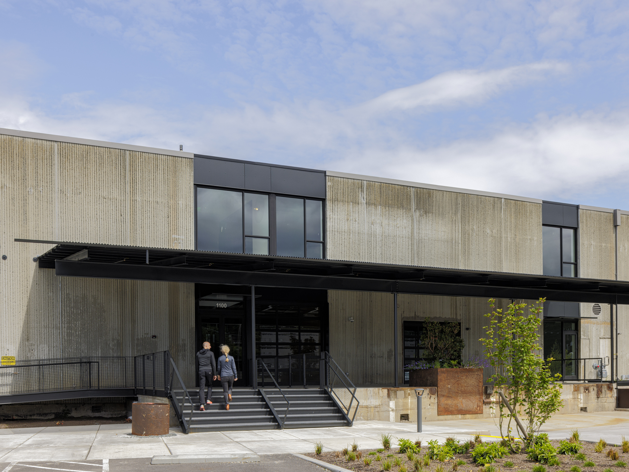 Front entrance with metal canopy, concrete steps, and weathered steel planters with two visitors ascending