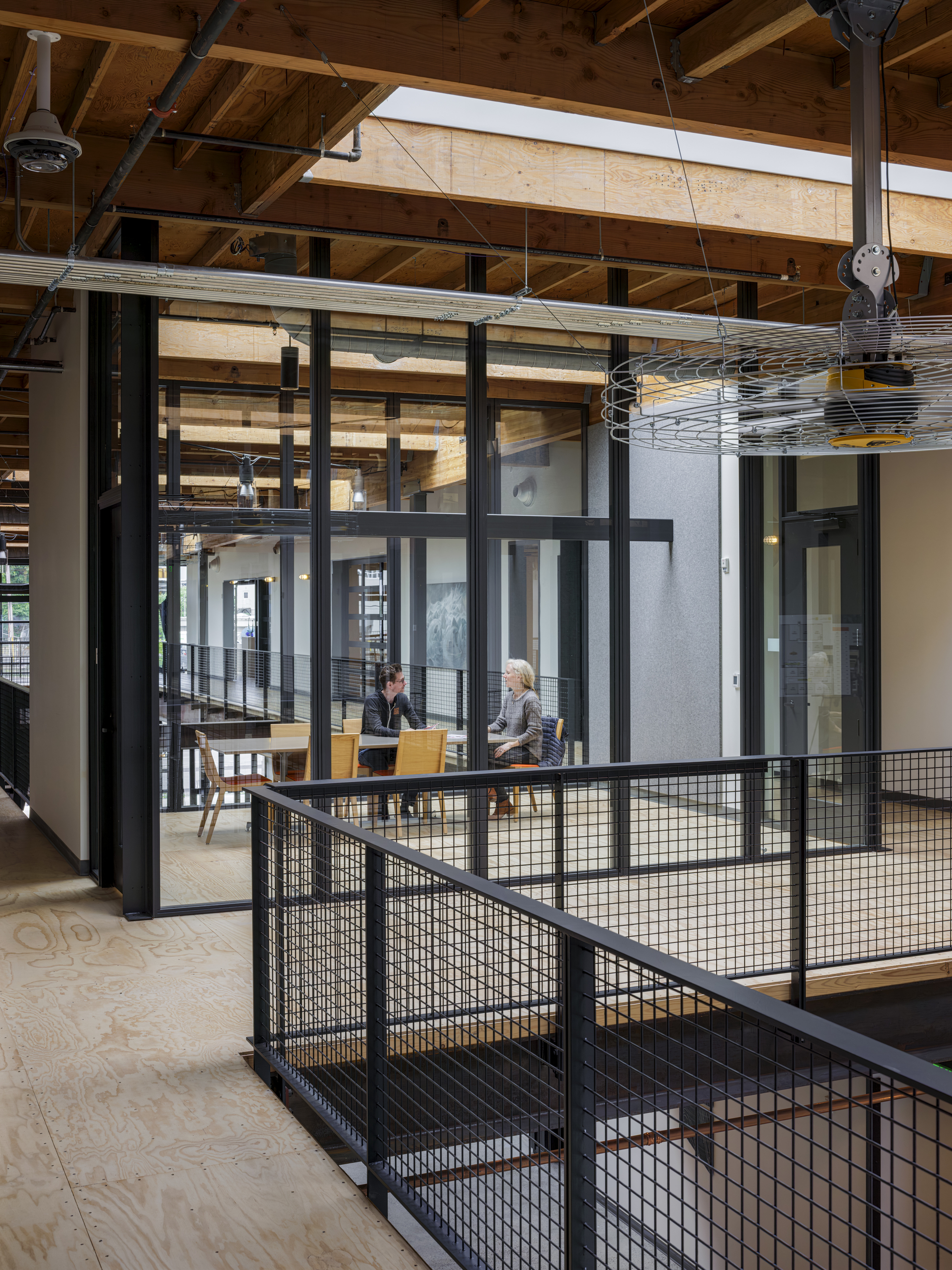 Double-height atrium with timber ceiling beams and black metal mesh stair railings overlooking a glazed meeting room