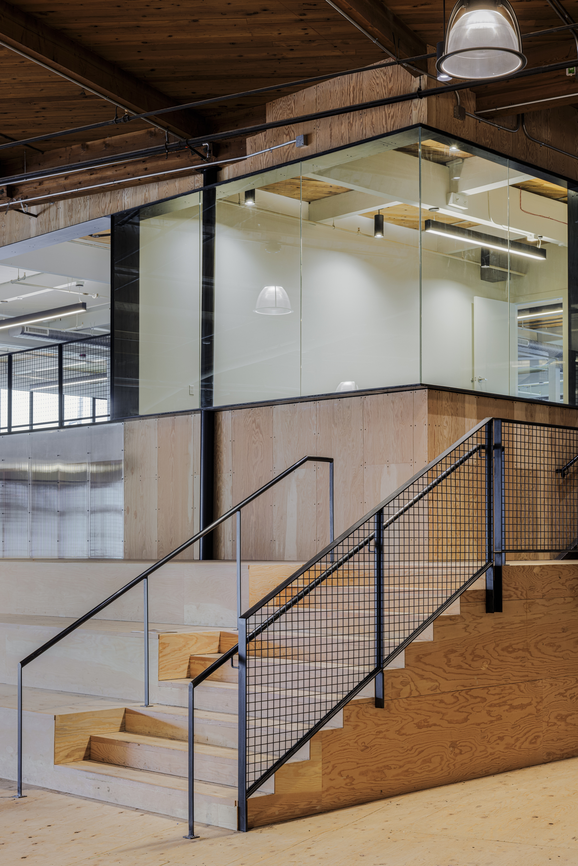 Interior staircase with plywood treads, black mesh railing and suspended glass conference room above