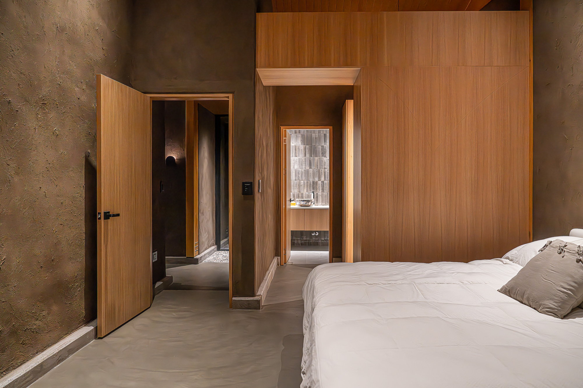 Bedroom with wood-paneled volume and polished concrete floor opening to ensuite bathroom through timber doorway