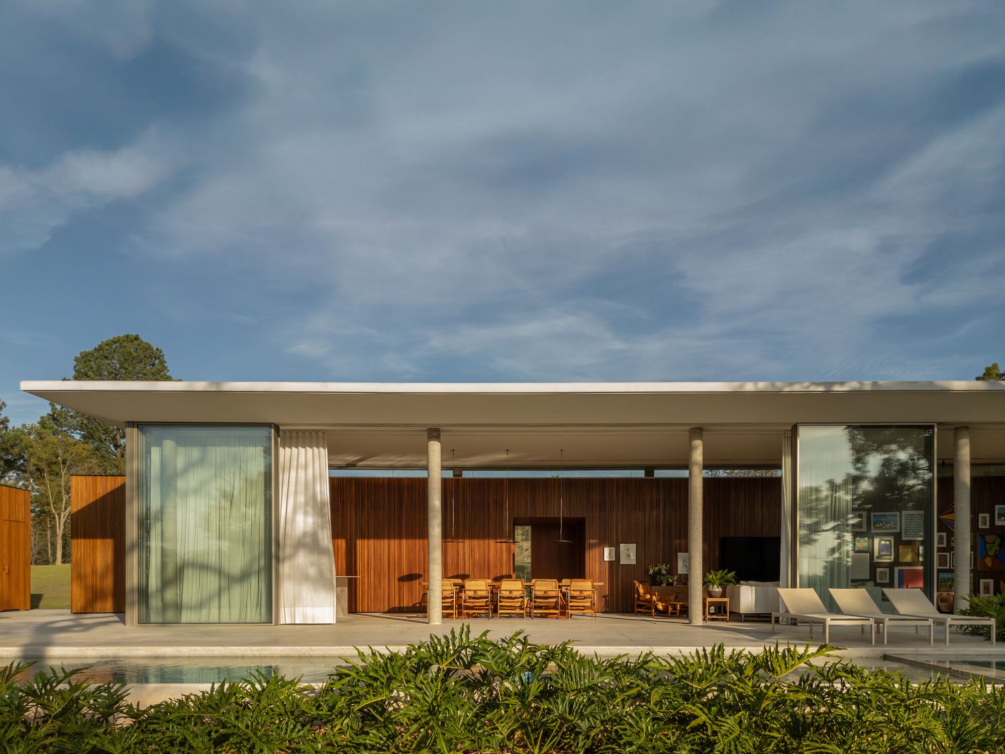 Covered poolside terrace with slender columns, timber-clad wall and recessed glass doors under afternoon light